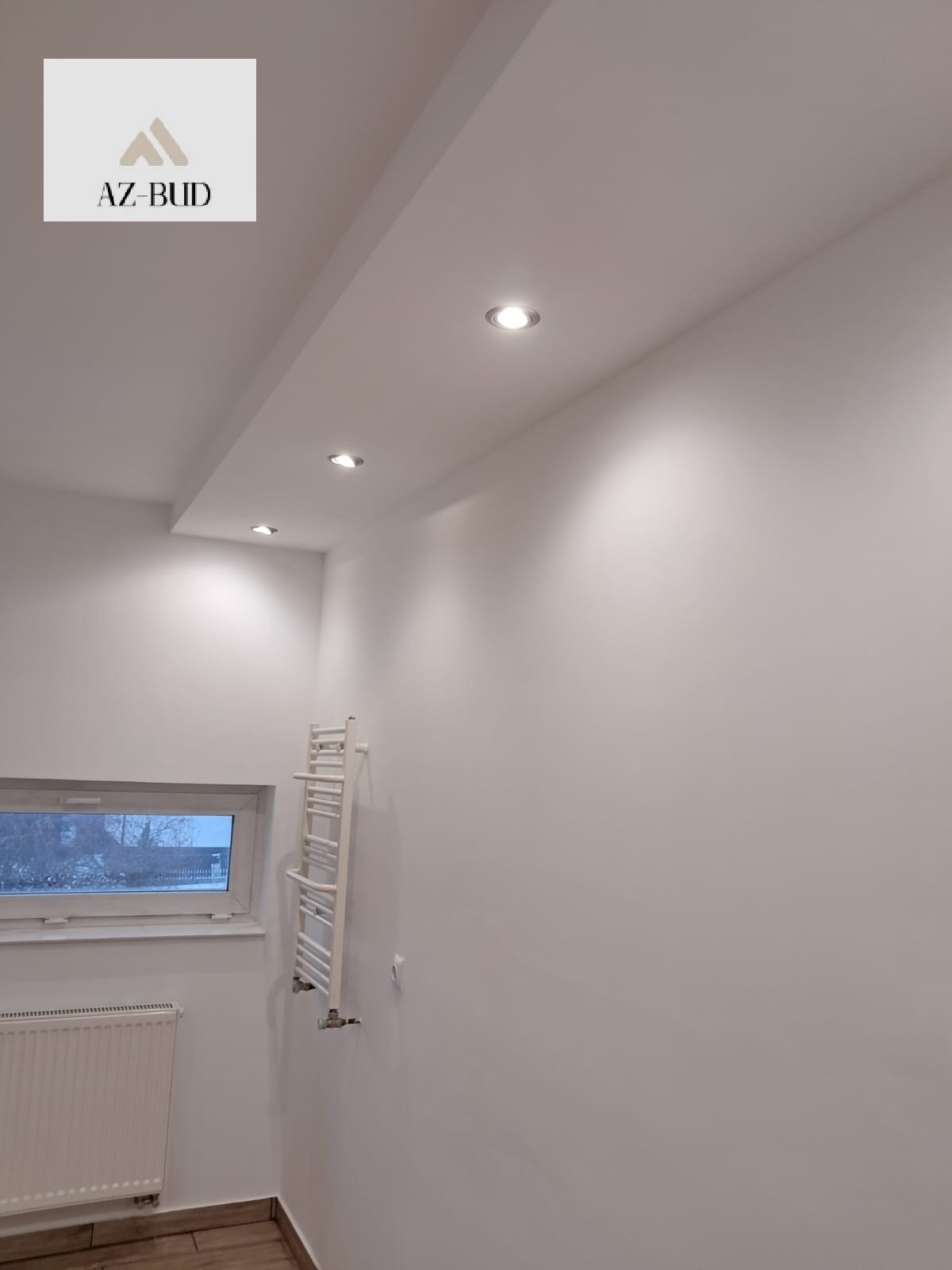 Białe, świeżo pomalowane wnętrze z grzejnikiem łazienkowym, widok na okno i trzy punktowe lampy sufitowe, logo firmy AZ-BUD w lewym górnym rogu.