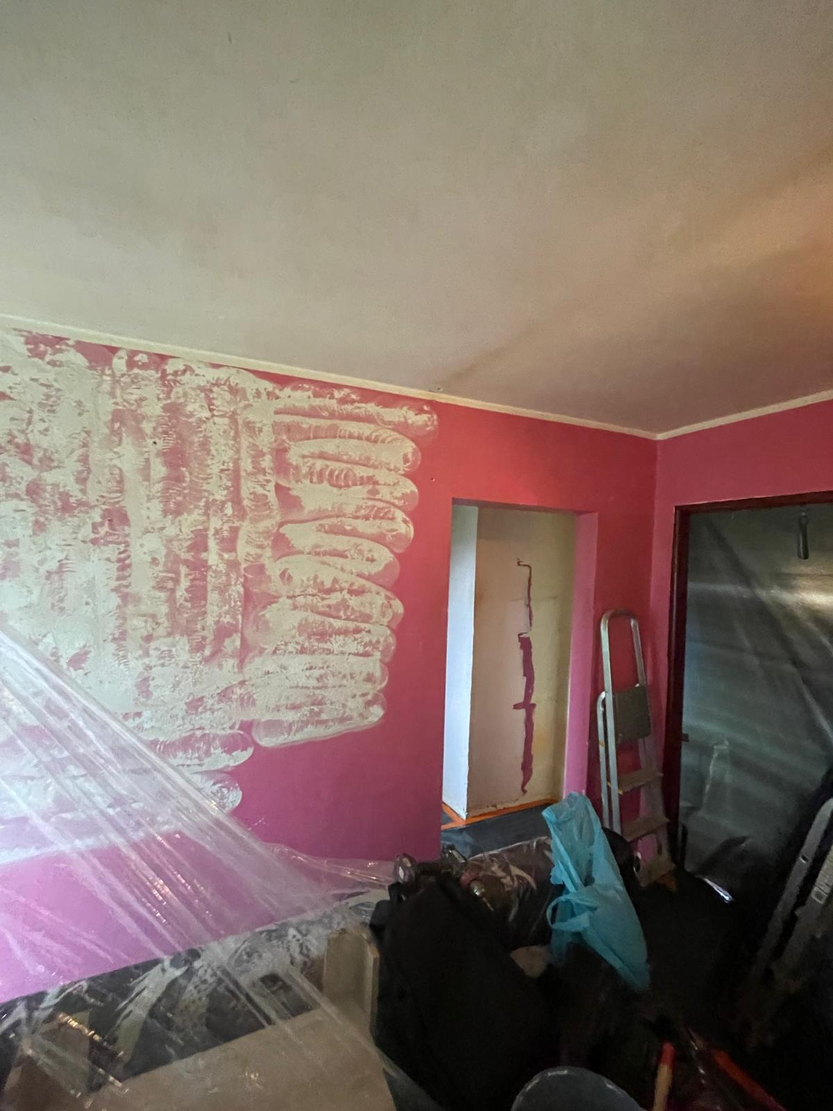 Świeżo nałożona warstwa gładzi szpachlowej na różowej ścianie w trakcie prac remontowych, widoczne folie ochronne i drabina