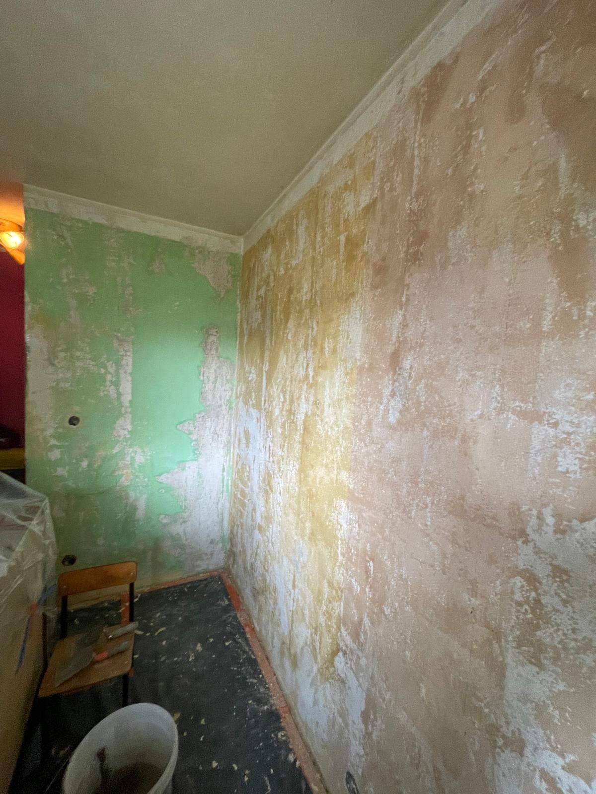 Pokój w trakcie remontu: ściany z resztkami starej farby i gładzi, widoczne warstwy zieleni i beżu, przygotowane do dalszych prac wykończeniowych, krzesło z narzędziami w rogu.