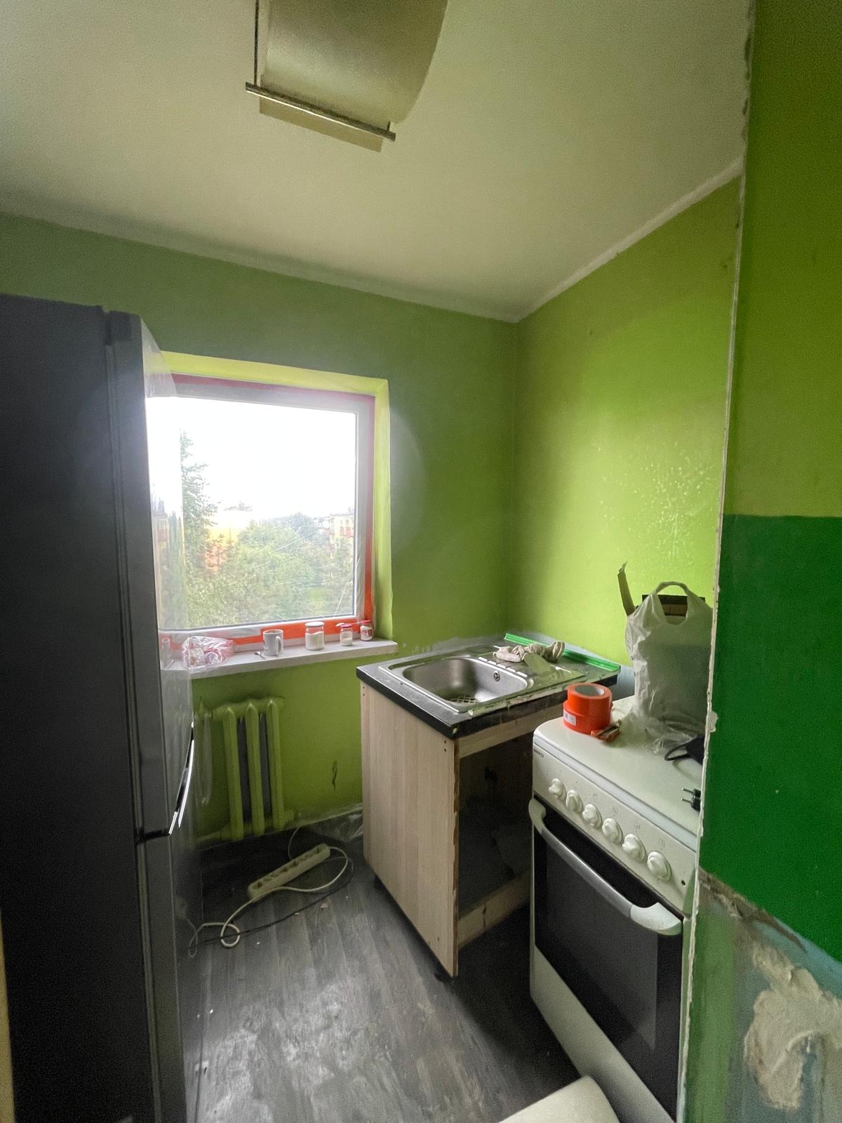 Kuchnia w trakcie remontu: świeżo pomalowane zielone ściany, okno zabezpieczone folią, surowa zabudowa pod zlewem, widoczne ślady prac malarskich i przygotowania do wykończenia.