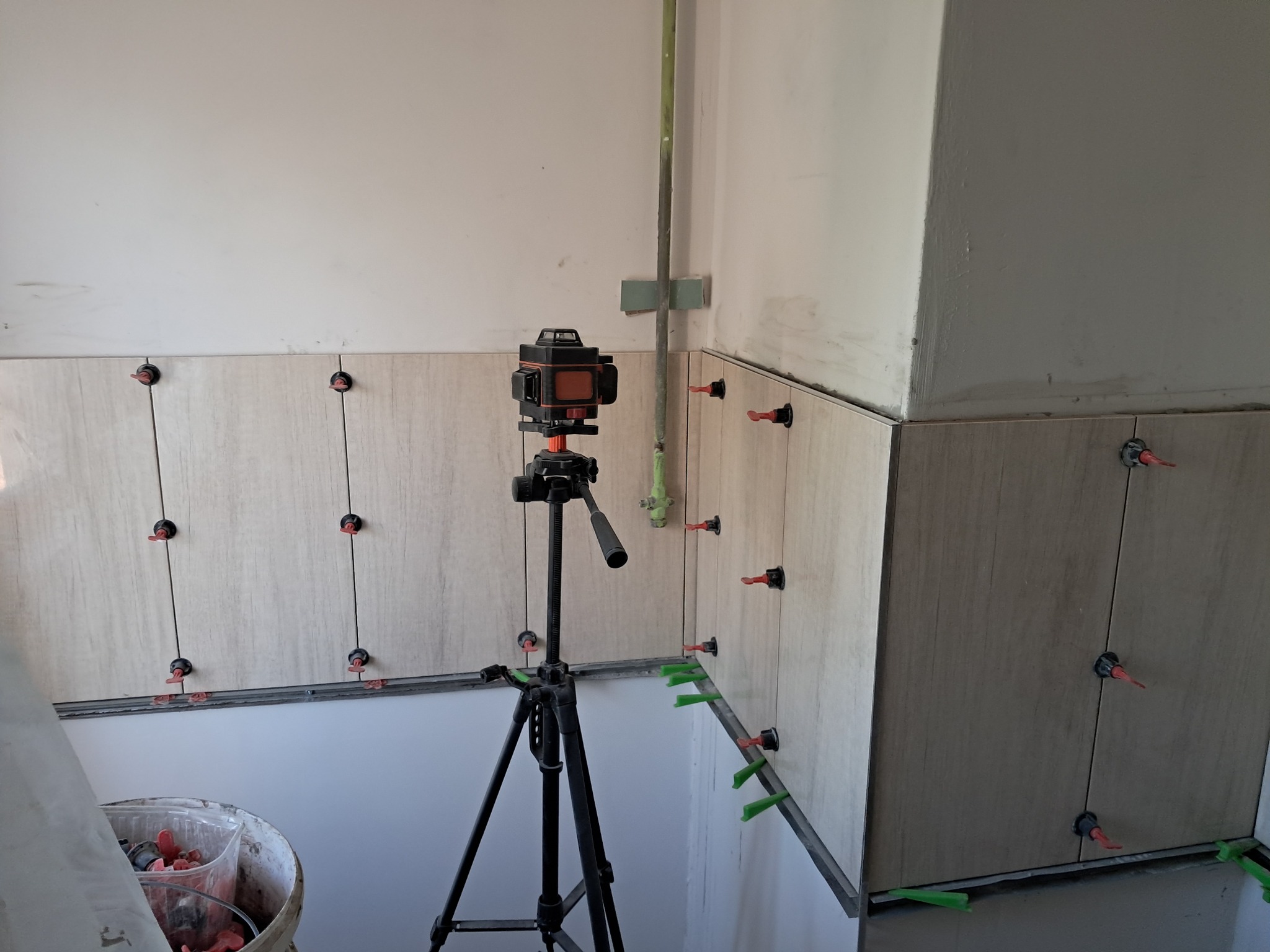 Ściana w trakcie układania płytek, widoczne narzędzia do poziomowania, laser krzyżowy na statywie, wiadro z elementami dystansowymi.