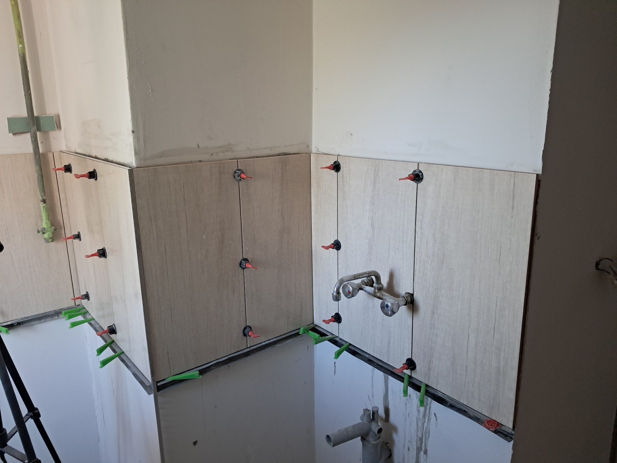 Świeżo położone, beżowe płytki ścienne w kuchni, z systemem poziomowania płytek (czerwone kliny i zielone dystanse) w trakcie montażu, widoczna bateria kuchenna.