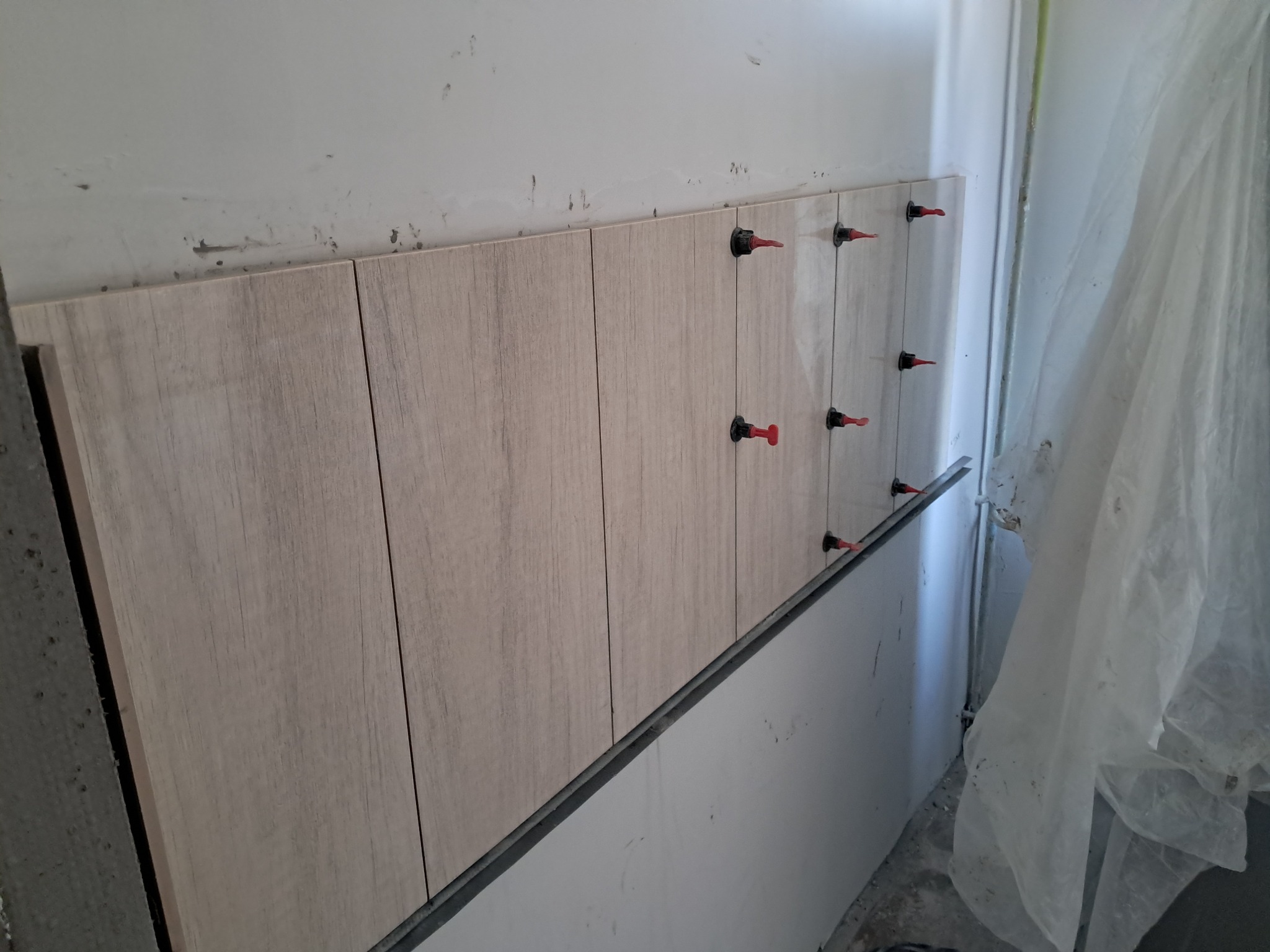 Świeżo ułożone, podłużne płytki imitujące drewno na ścianie, z użyciem systemu poziomowania z czerwonymi klinami, widoczne ślady po kleju na górze ściany.