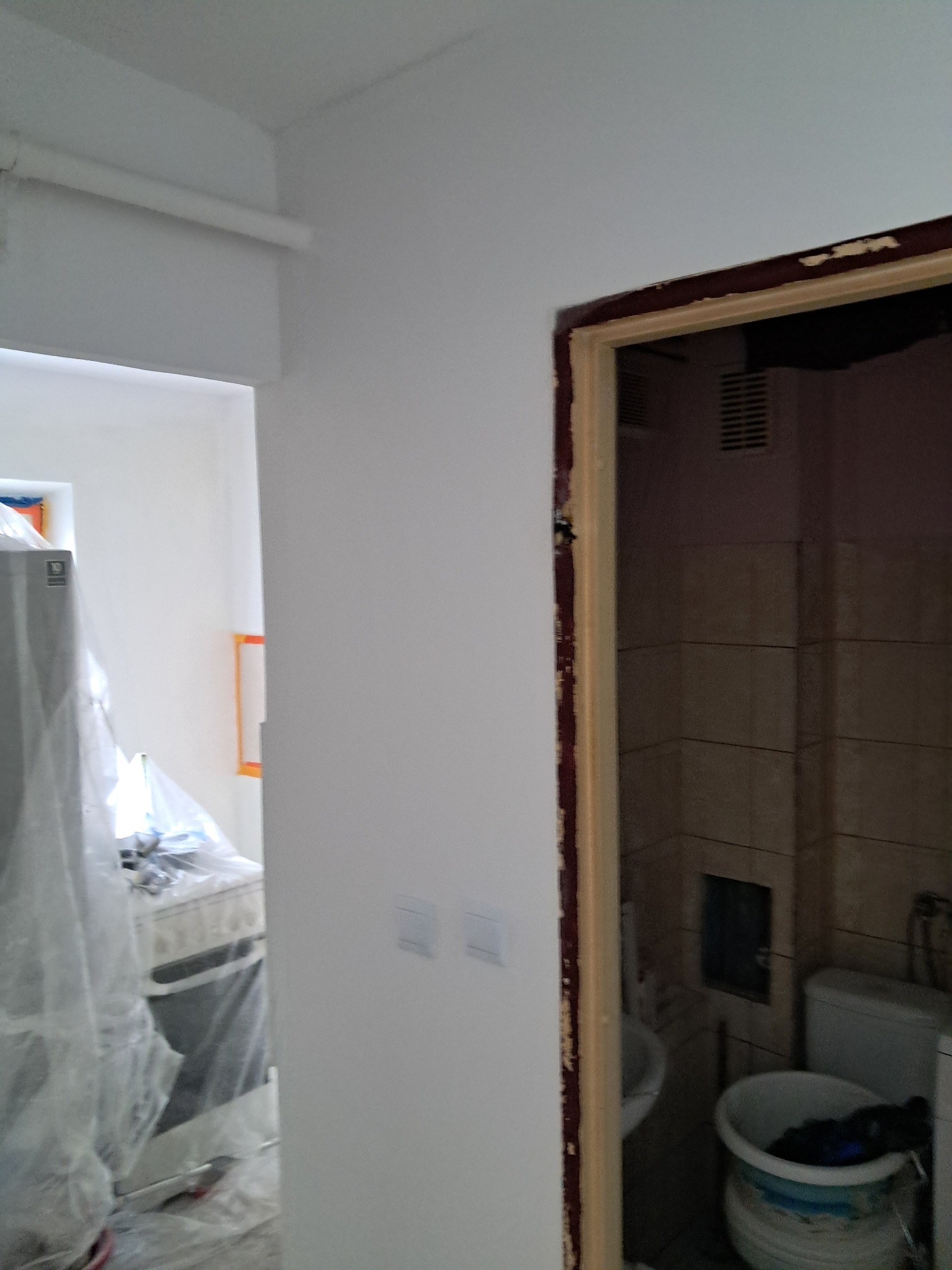 Świeżo pomalowane na biało ściany mieszkania w trakcie remontu, widoczne zabezpieczone folią meble w sąsiednim pomieszczeniu oraz wejście do łazienki z widocznymi starymi płytkami.