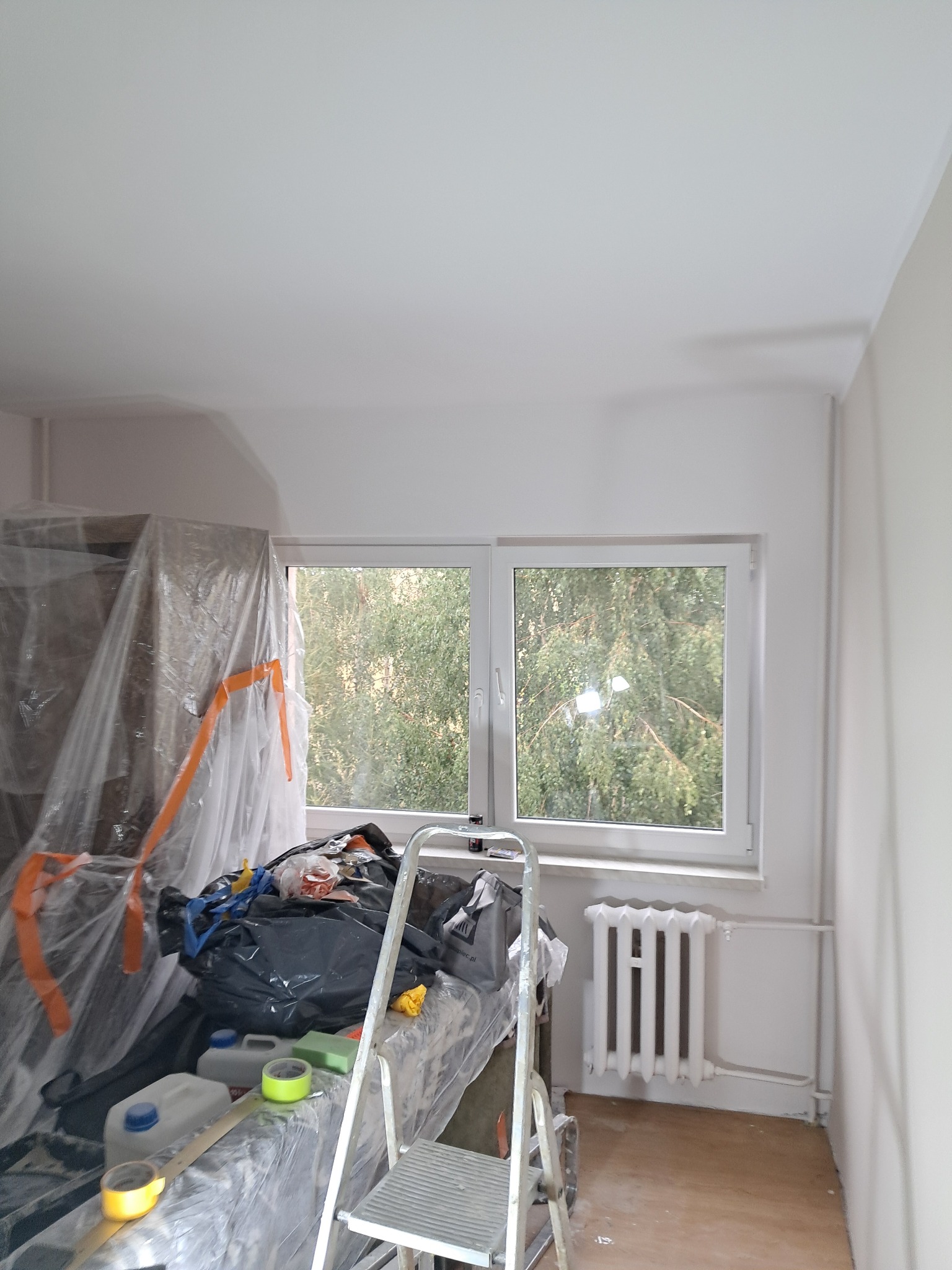 Pokój w trakcie malowania: zabezpieczone folią meble, drabina, widok na drzewa za oknem, świeżo pomalowane ściany i sufit.
