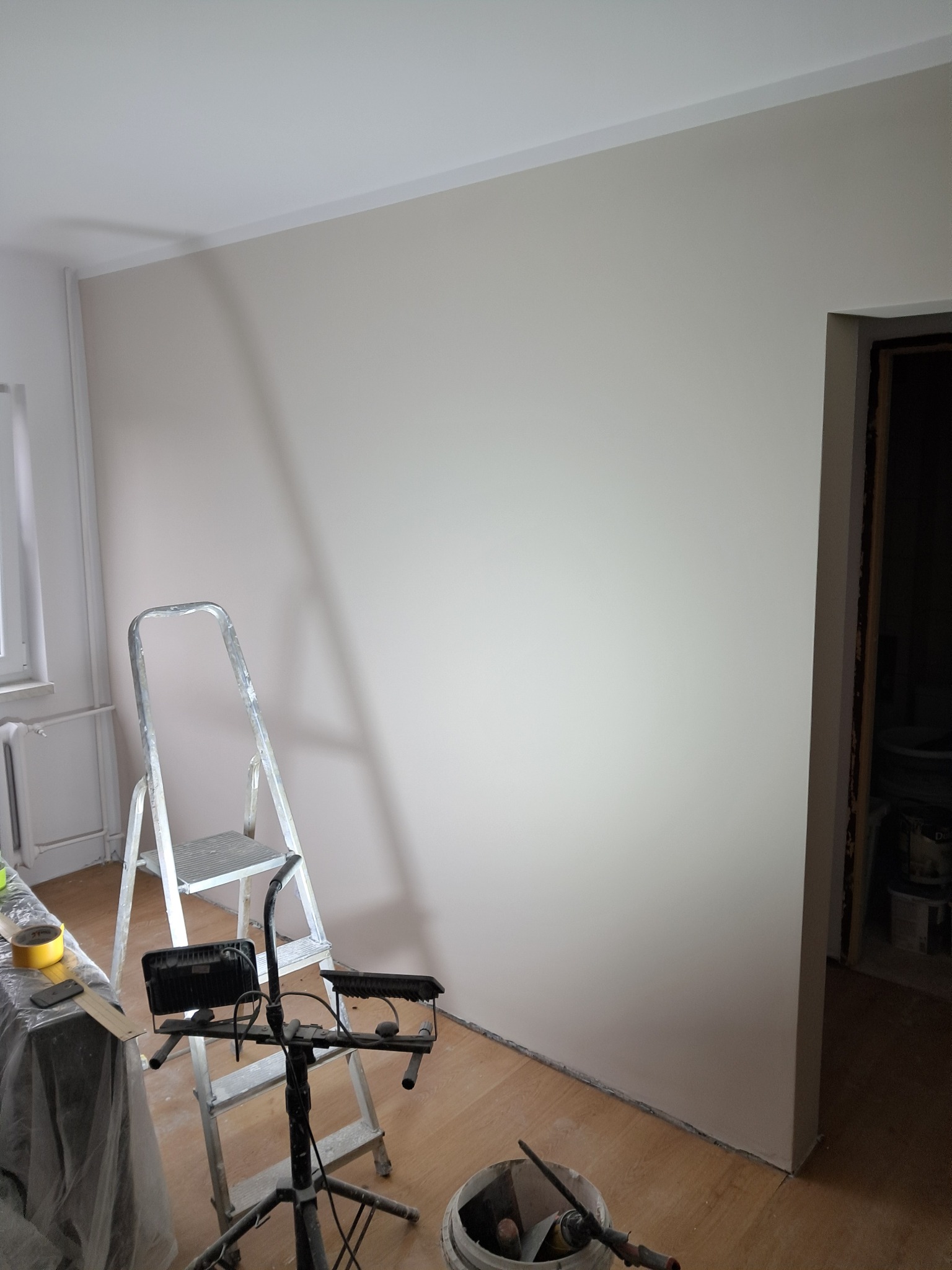 Świeżo pomalowana ściana w odcieniach beżu, widoczna drabina malarska, oświetlenie budowlane, wiadro z narzędziami malarskimi i częściowo zasłonięte okno.