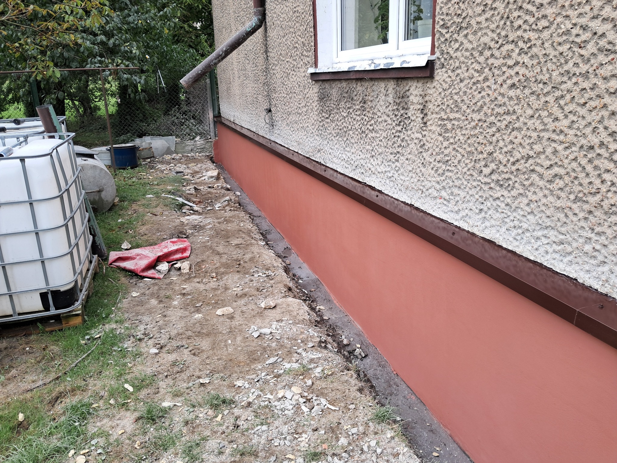 Dolna część elewacji budynku po nałożeniu nowej, gładkiej, brązowo-czerwonej warstwy tynku, kontrastująca z chropowatą, szarą strukturą powyżej, widoczne ślady prac wokół.