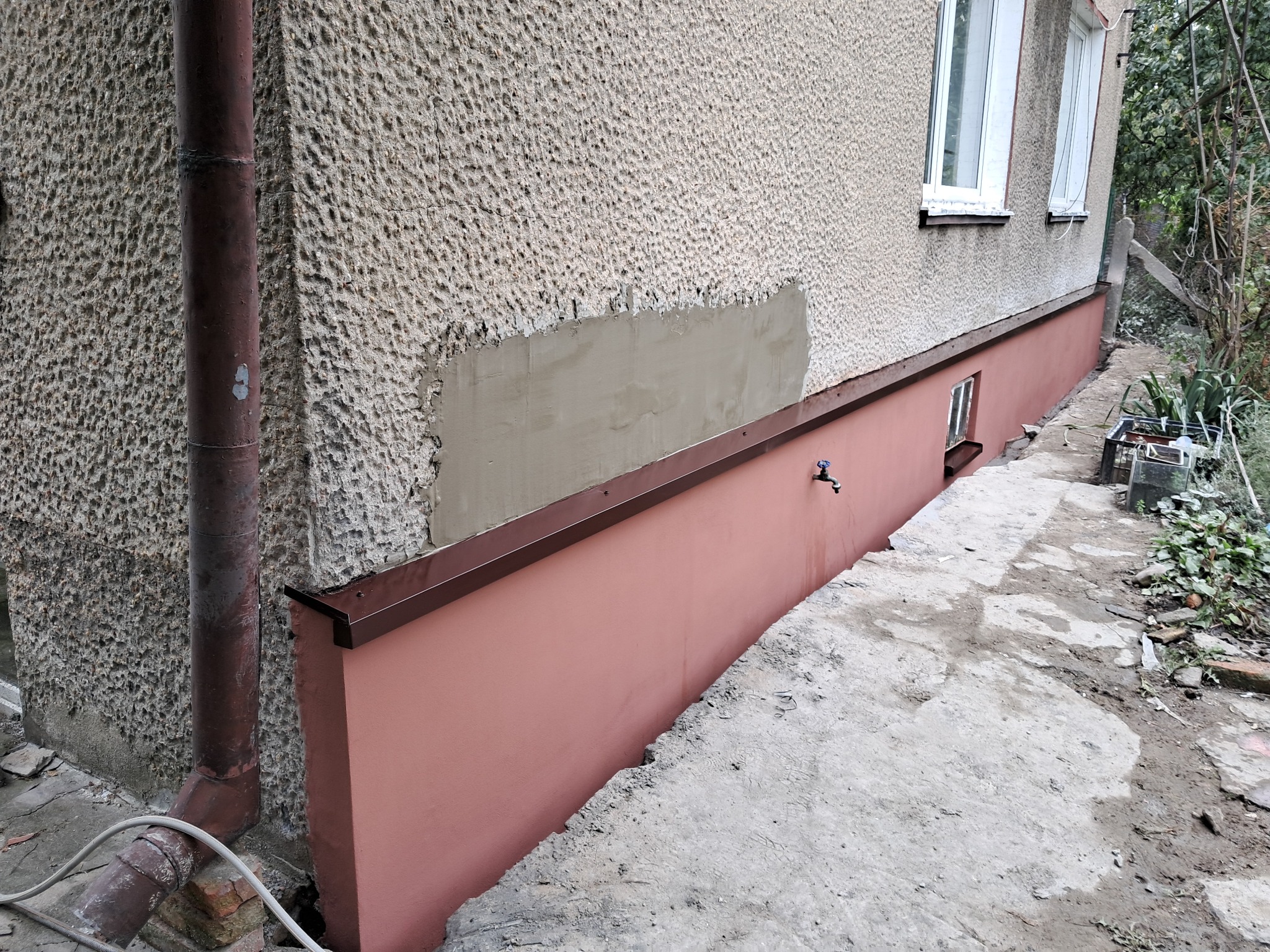 Fragment ściany budynku z widocznym fragmentem nowej gładzi na chropowatej elewacji, pomalowany cokół na różowo, rura spustowa z widocznymi śladami korozji.
