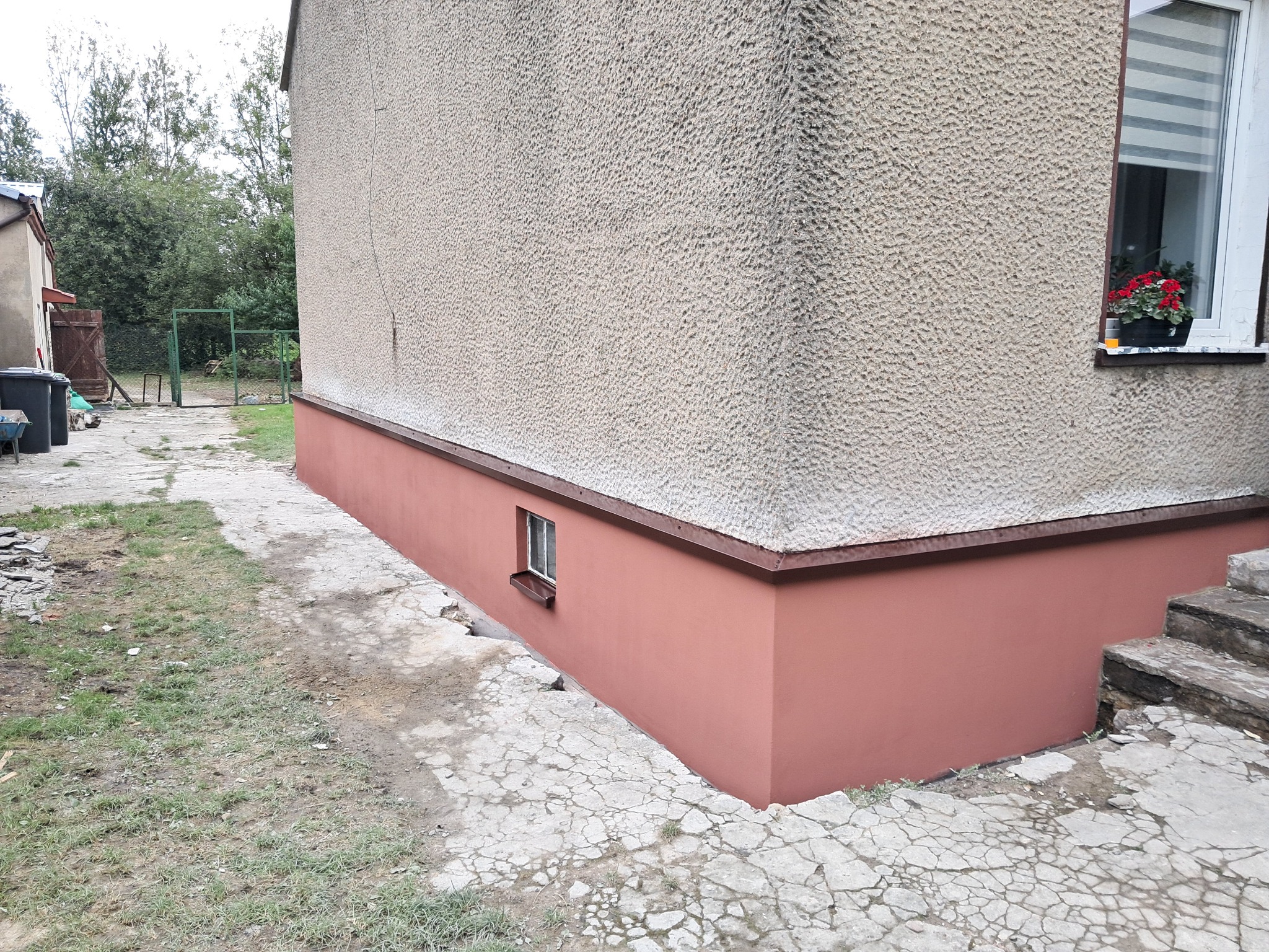 Świeżo pomalowana, dolna część elewacji budynku w kolorze ceglastym, kontrastująca z szarą, chropowatą górną częścią ściany i betonowym chodnikiem z widocznymi pęknięciami.
