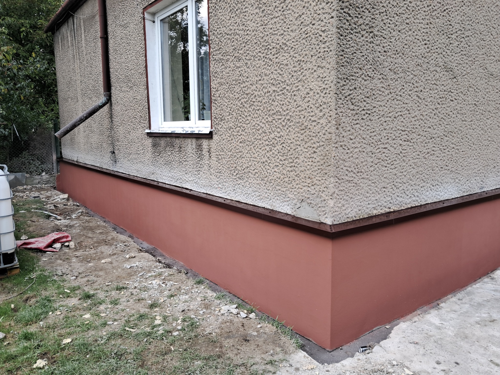 Odnowiona elewacja budynku z pomalowanym cokołem na kolor ceglasty, widoczne okno z białą ramą i fragment ogrodu z zielenią.