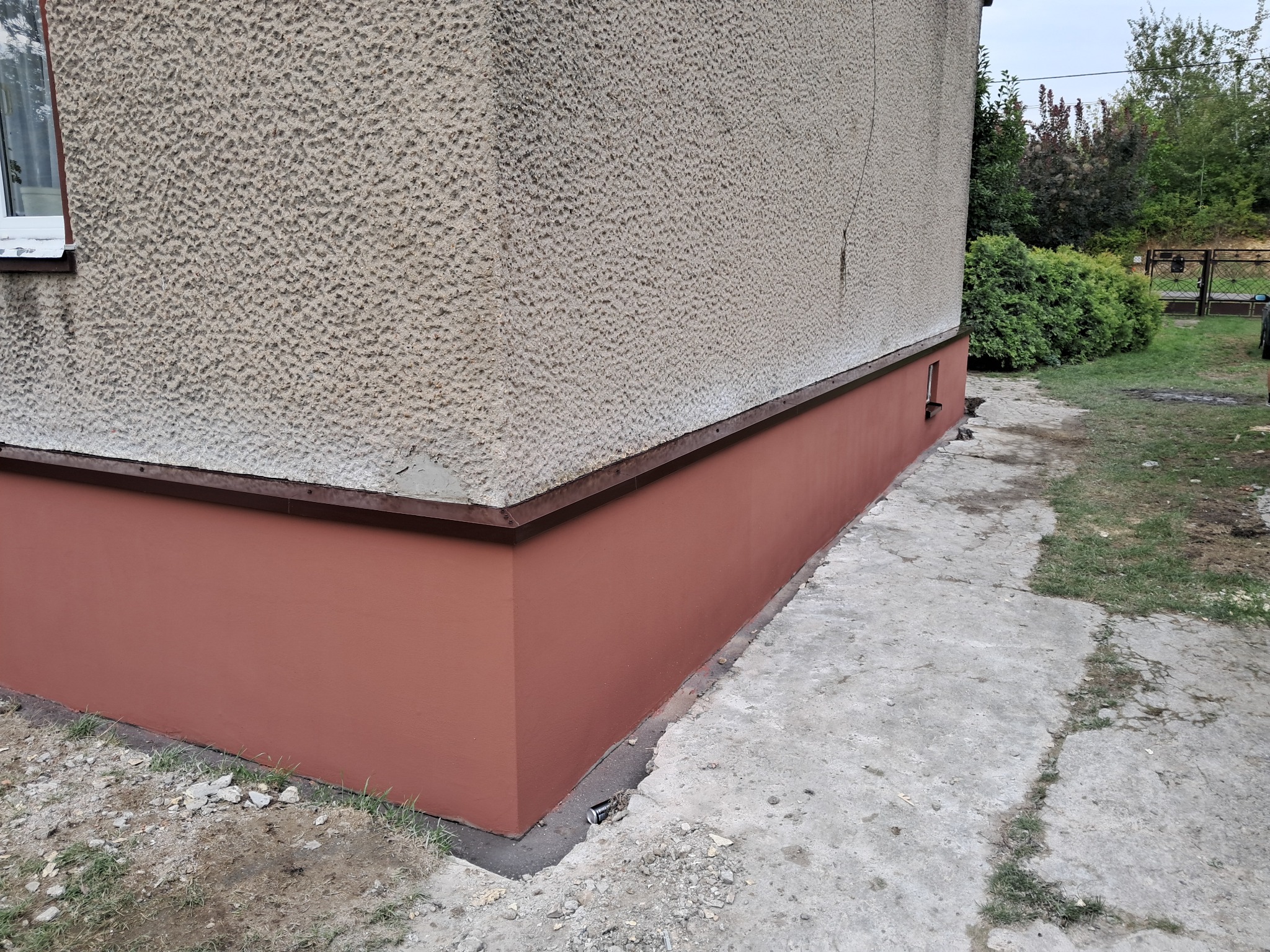 Fragment ściany budynku z widocznym odnowionym cokołem w kolorze terakoty, szara elewacja z fakturą baranka oraz betonowy chodnik z chwastami.