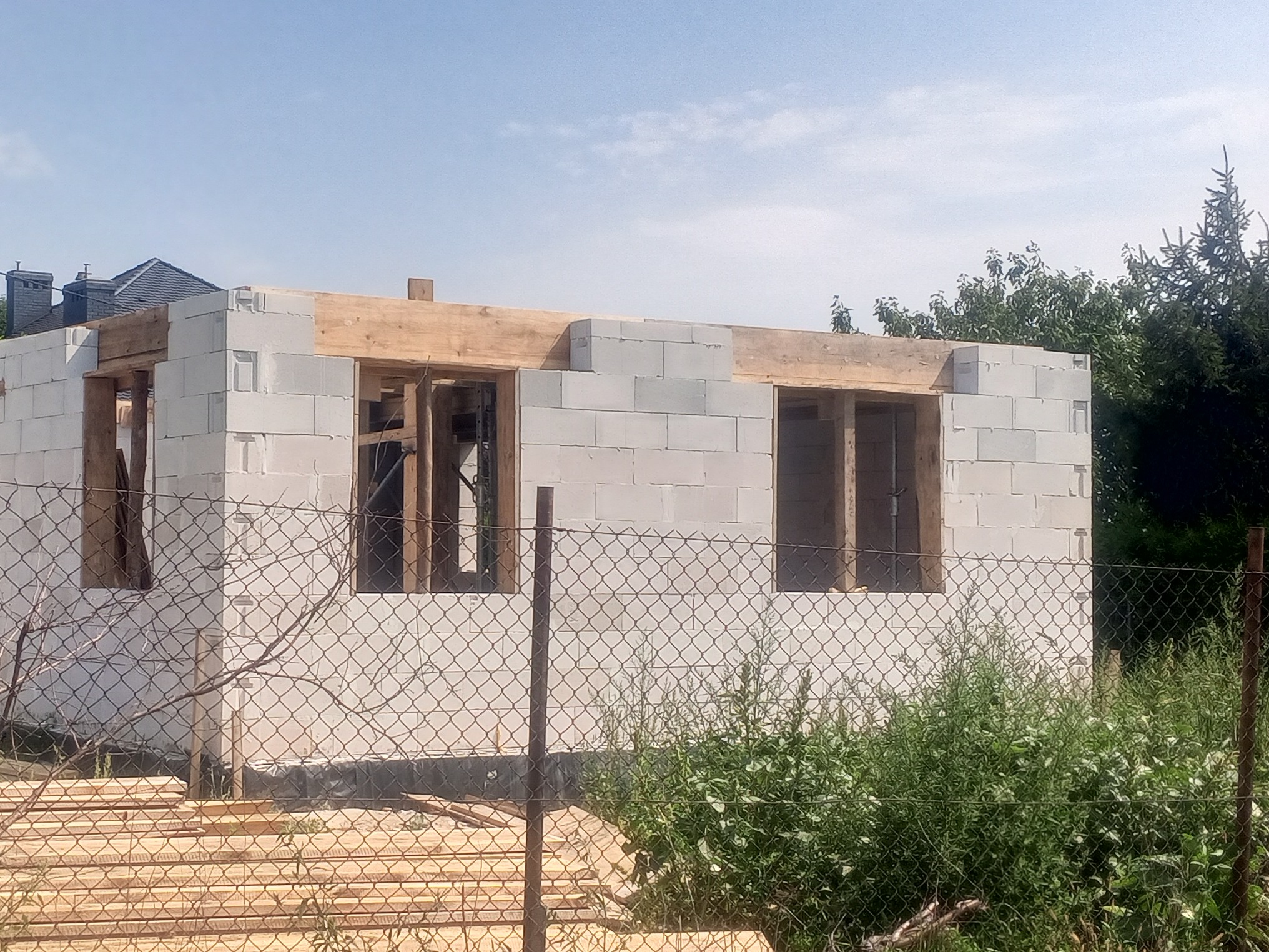 Wznoszenie ścian parteru domu z bloczków betonu komórkowego, widoczne otwory okienne z drewnianymi nadprożami, teren budowy zabezpieczony siatką ogrodzeniową.