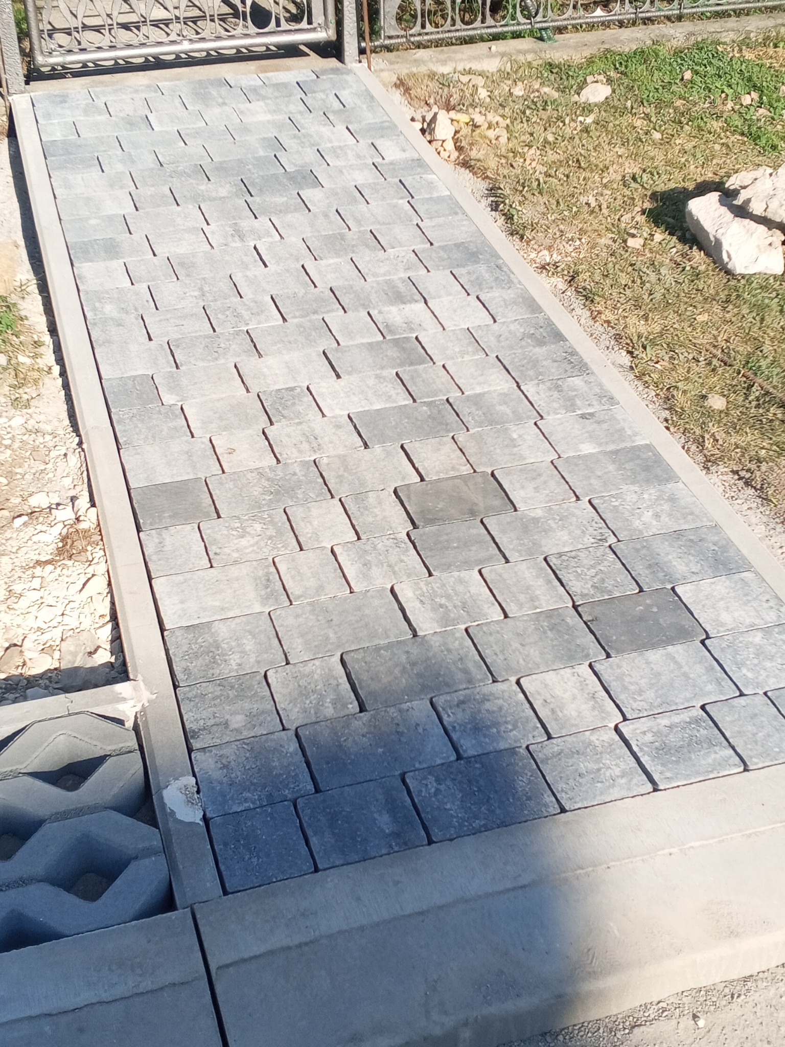 Ścieżka z szarej kostki brukowej ułożonej w prostokątnym wzorze, obramowana betonowym krawężnikiem, prowadząca do metalowej bramki; widoczne fragmenty trawy i ziemi.