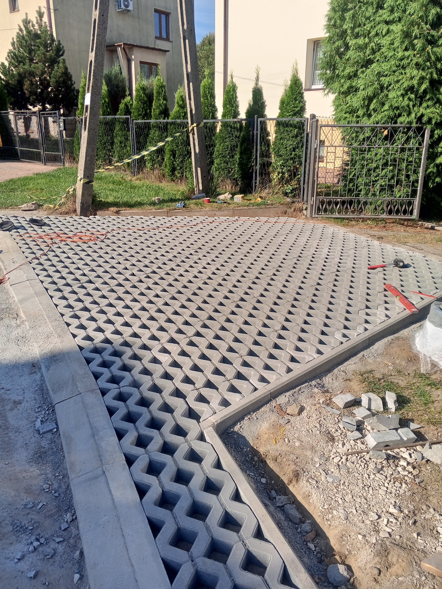 Szara ażurowa kostka brukowa ułożona na podjeździe, widoczne narzędzia brukarskie i fragmenty kostki do docięcia, w tle brama i budynek.