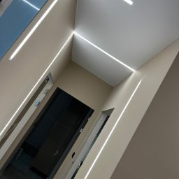 T&S Remonty i wykończenia - Korytarz z beżowymi ścianami i sufitem, oświetlony liniowymi lampami LED tworzącymi geometryczny wzór na suficie i ścianach, widoczne drzwi do pomieszczeń.