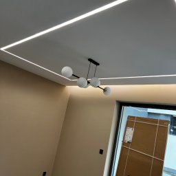 T&S Remonty i wykończenia - Nowoczesne oświetlenie sufitowe LED w kształcie prostokąta, centralnie umieszczony żyrandol z kulistymi kloszami, ściany pomalowane na beżowo, widoczne kartony przy oknie.