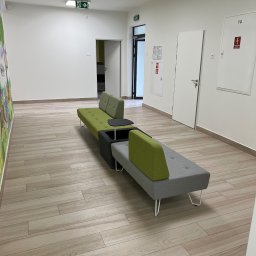 T&S Remonty i wykończenia - Długi, modułowy zestaw wypoczynkowy w odcieniach szarości i zieleni w przestronnym korytarzu z panelami podłogowymi imitującymi drewno, białe ściany z elementami graficznymi i drzwiami.