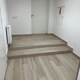T&S Remonty i wykończenia - Wykończone schody wejściowe do budynku, pokryte płytkami imitującymi drewno, z widocznym grzejnikiem i instalacją przeciwpożarową na białej ścianie.