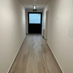 Korytarz z nowymi płytkami podłogowymi imitującymi drewno, świeżo pomalowane ściany w odcieniach bieli, czarne drzwi na końcu korytarza z widocznym znakiem ewakuacyjnym.