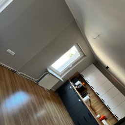 T&S Remonty i wykończenia - Szare, świeżo pomalowane poddasze z oknem dachowym, widoczne gładkie ściany, grzejnik, instalacja elektryczna, drewnopodobna podłoga oraz zabudowa kuchenna z czarnymi i białymi frontami.