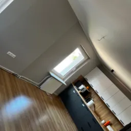 Szare, świeżo pomalowane poddasze z oknem dachowym, widoczne gładkie ściany, grzejnik, instalacja elektryczna, drewnopodobna podłoga oraz zabudowa kuchenna z czarnymi i białymi frontami.