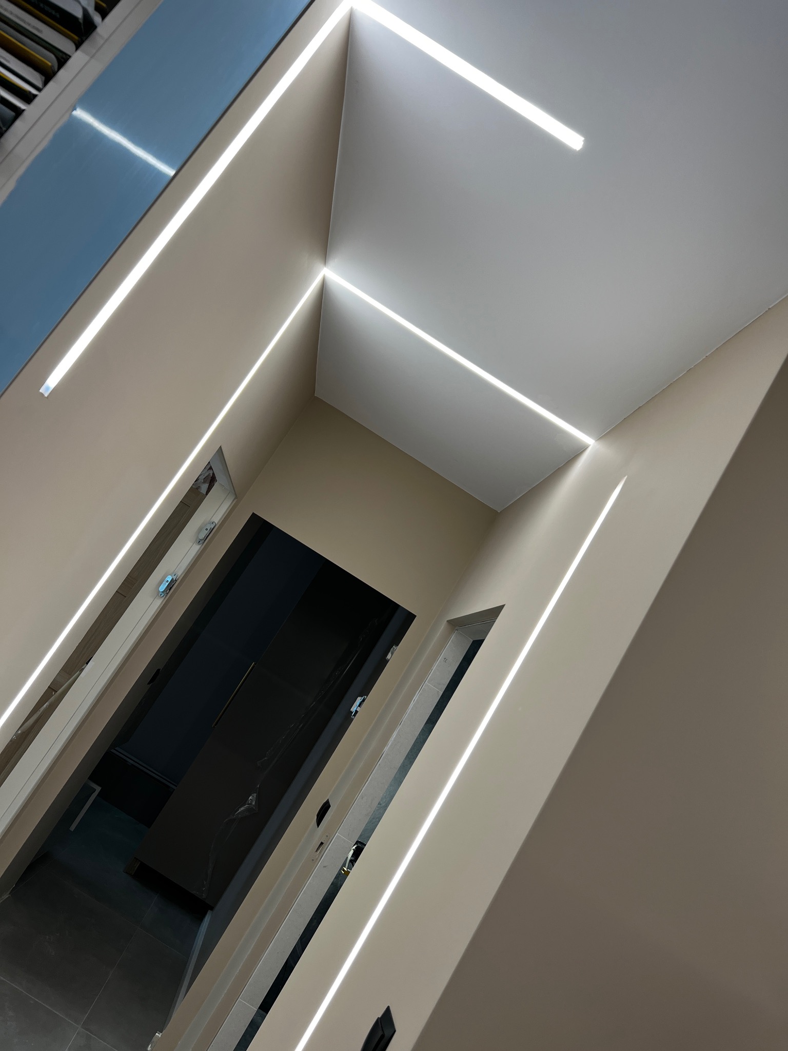 Korytarz z beżowymi ścianami i sufitem, oświetlony liniowymi lampami LED tworzącymi geometryczny wzór na suficie i ścianach, widoczne drzwi do pomieszczeń.