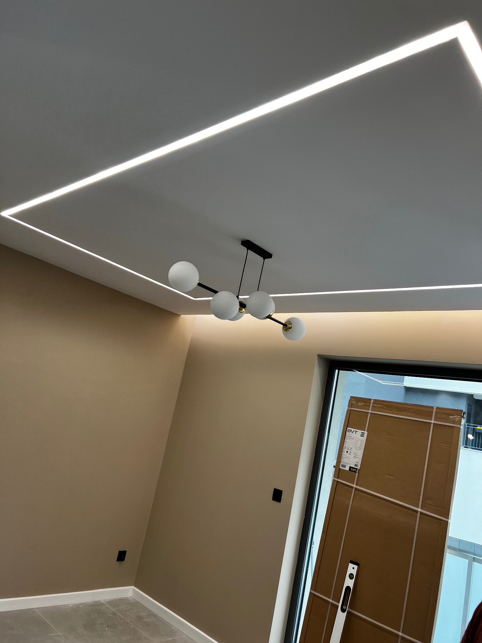 Nowoczesne oświetlenie sufitowe LED w kształcie prostokąta, centralnie umieszczony żyrandol z kulistymi kloszami, ściany pomalowane na beżowo, widoczne kartony przy oknie.