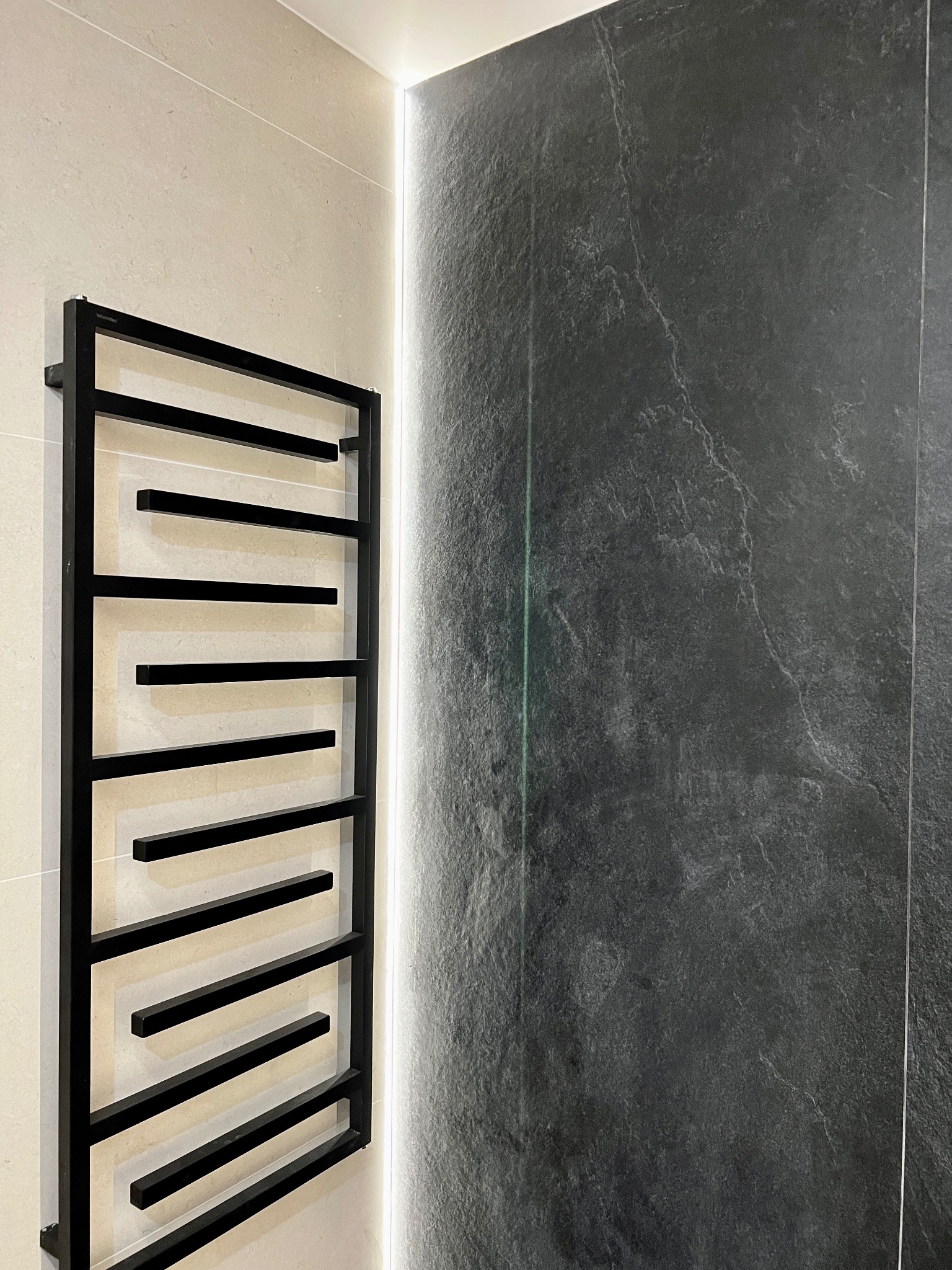 Nowoczesna łazienka z czarnym grzejnikiem drabinkowym na tle beżowych płytek i ściany z szarym kamieniem, oświetlona pionową listwą LED.
