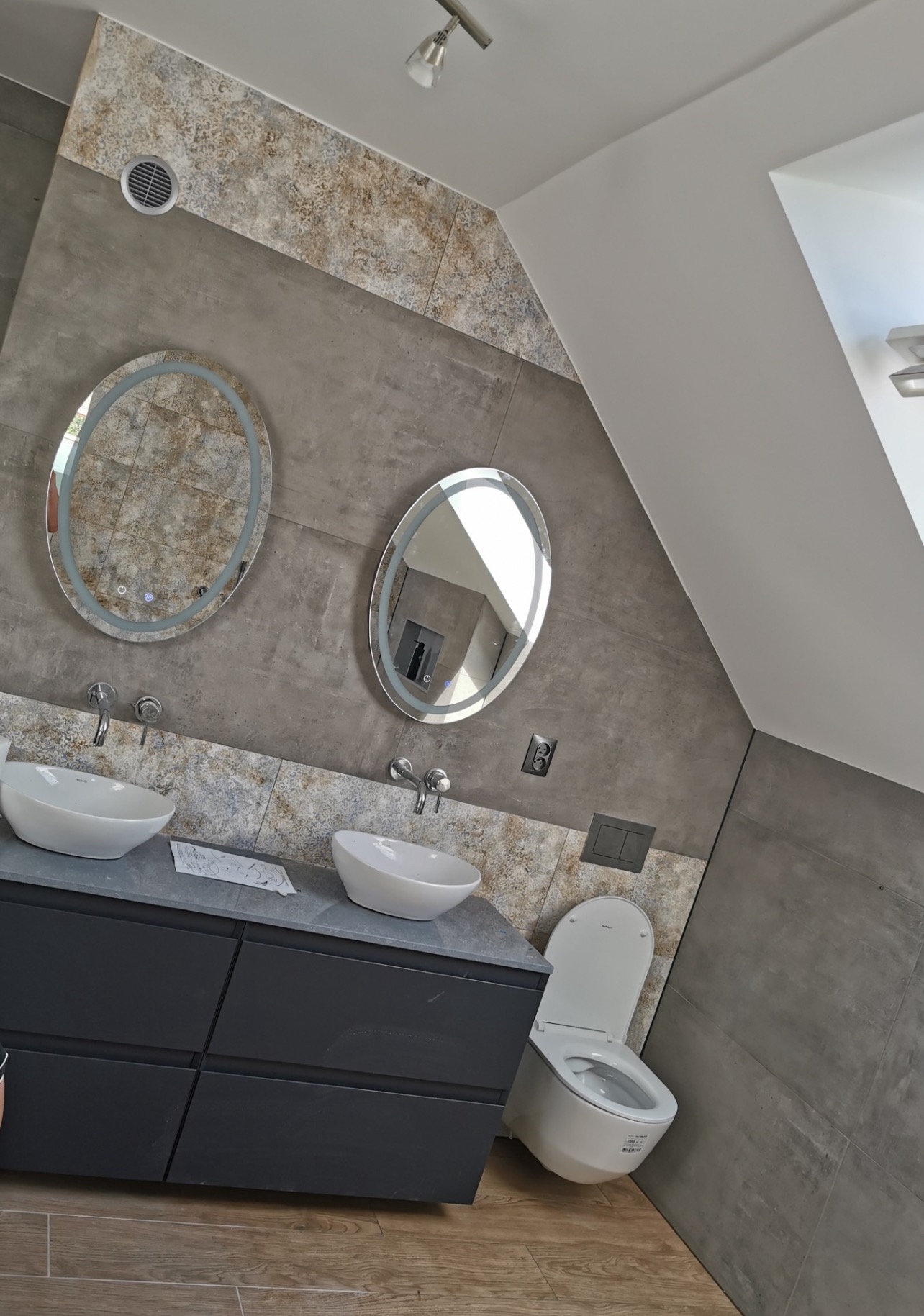 Nowoczesna łazienka z dwoma okrągłymi lustrami z podświetleniem LED, dwiema umywalkami nablatowymi na szafce z szufladami w kolorze grafitowym, wiszącą miską WC i szarą mozaiką na ścianach.
