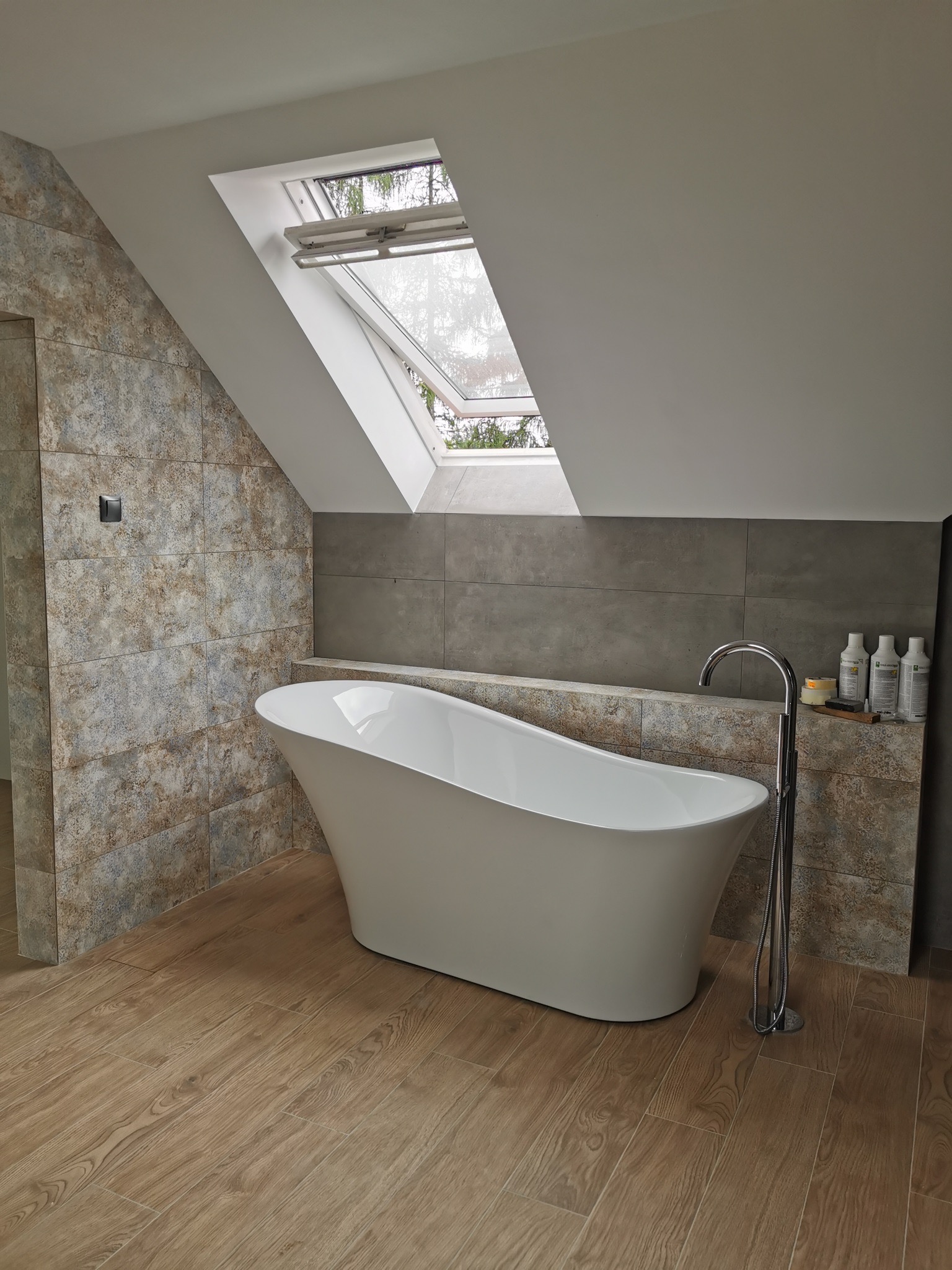 Nowoczesna łazienka na poddaszu z wolnostojącą wanną, drewnopodobną podłogą i płytkami w dwóch odcieniach szarości, doświetlona oknem dachowym.