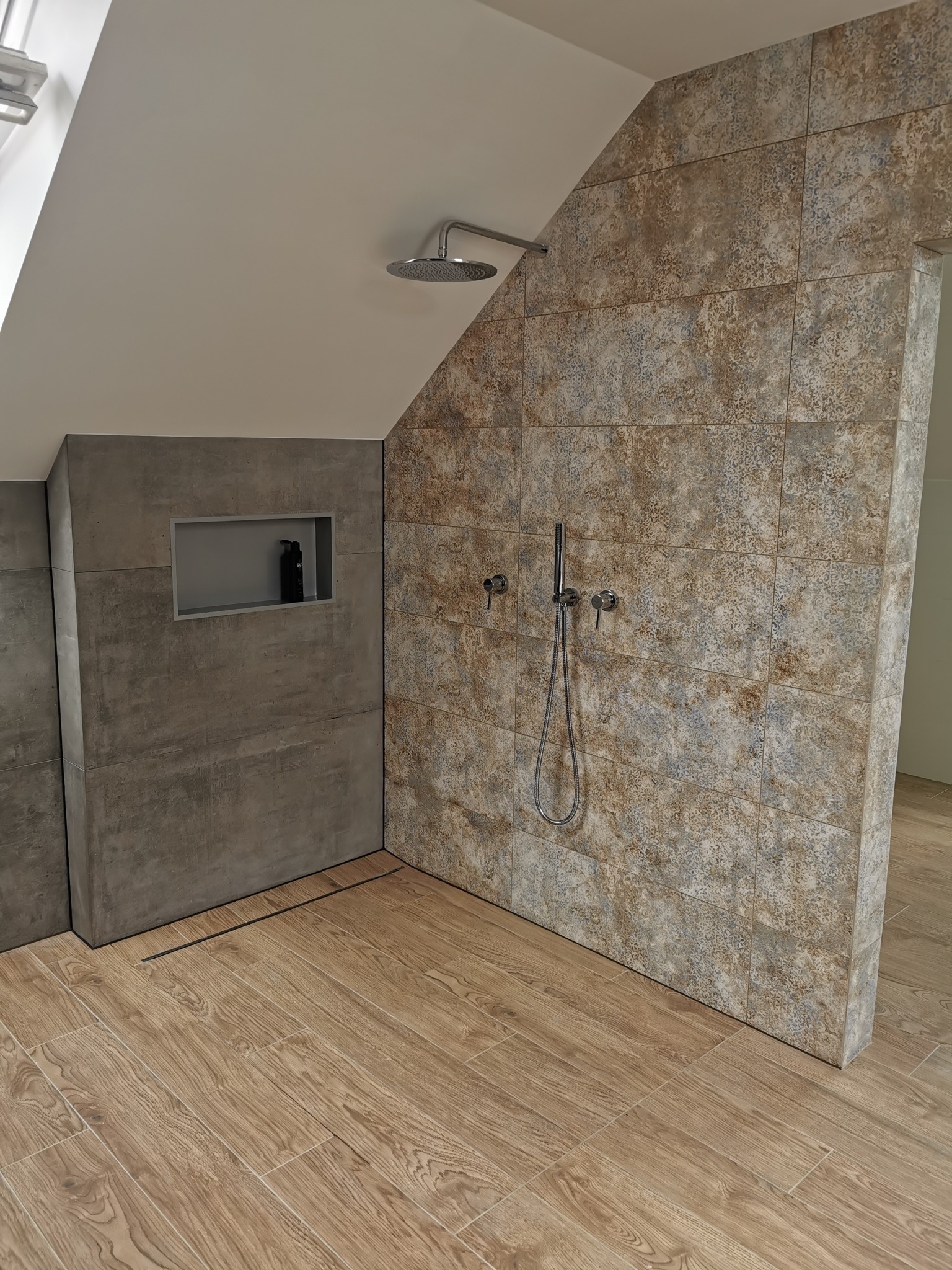 Nowoczesna kabina prysznicowa w skosie poddasza, wyłożona płytkami imitującymi kamień w odcieniach beżu i szarości, z wnęką na kosmetyki i odpływem liniowym.