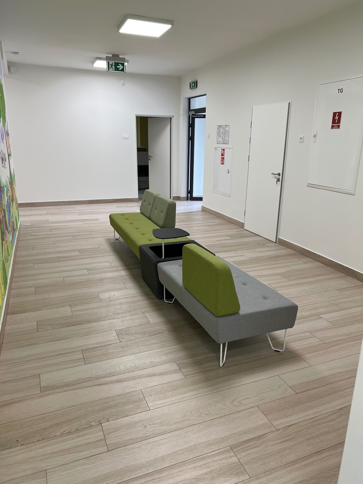 Długi, modułowy zestaw wypoczynkowy w odcieniach szarości i zieleni w przestronnym korytarzu z panelami podłogowymi imitującymi drewno, białe ściany z elementami graficznymi i drzwiami.