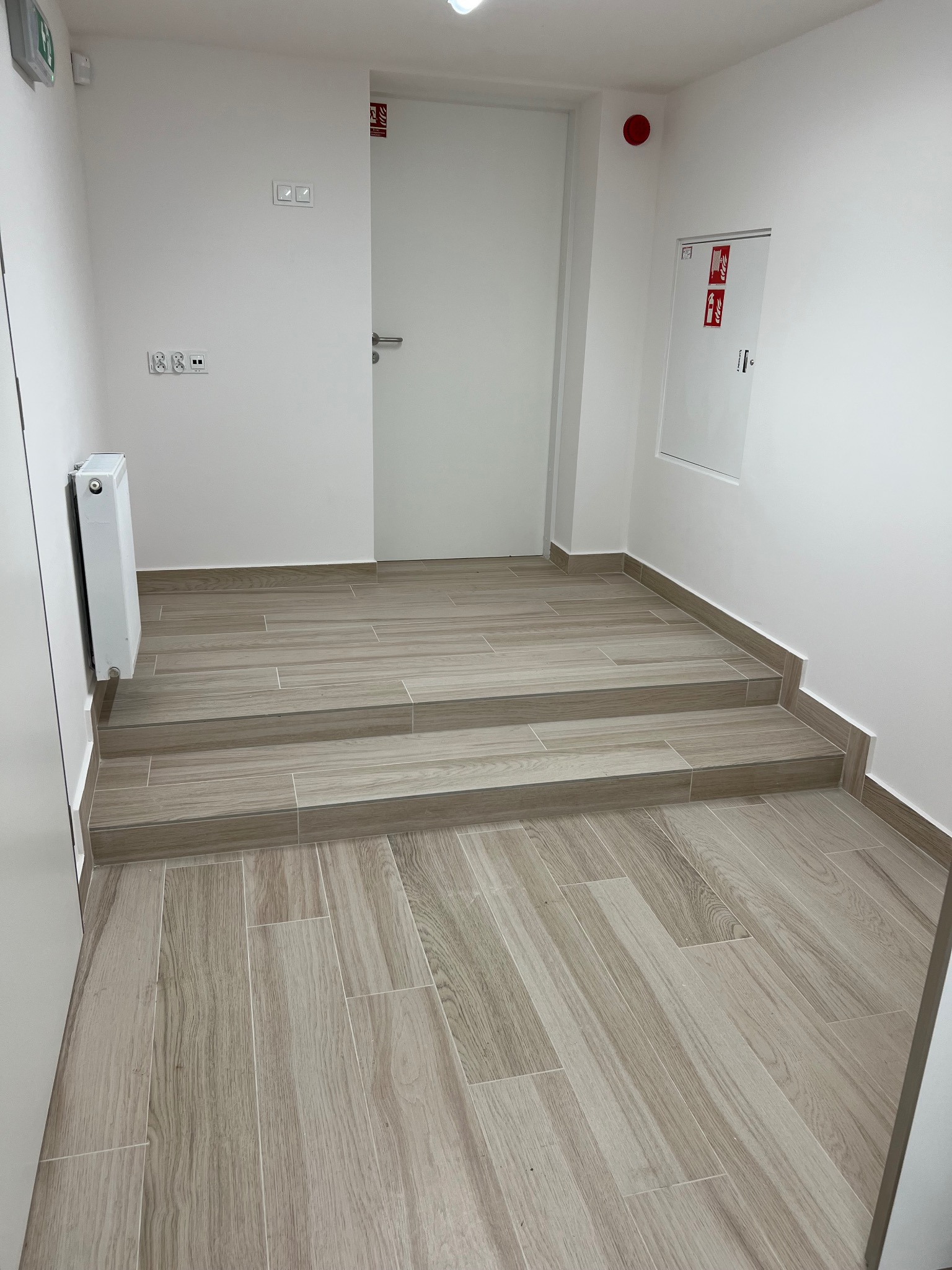 Wykończone schody wejściowe do budynku, pokryte płytkami imitującymi drewno, z widocznym grzejnikiem i instalacją przeciwpożarową na białej ścianie.