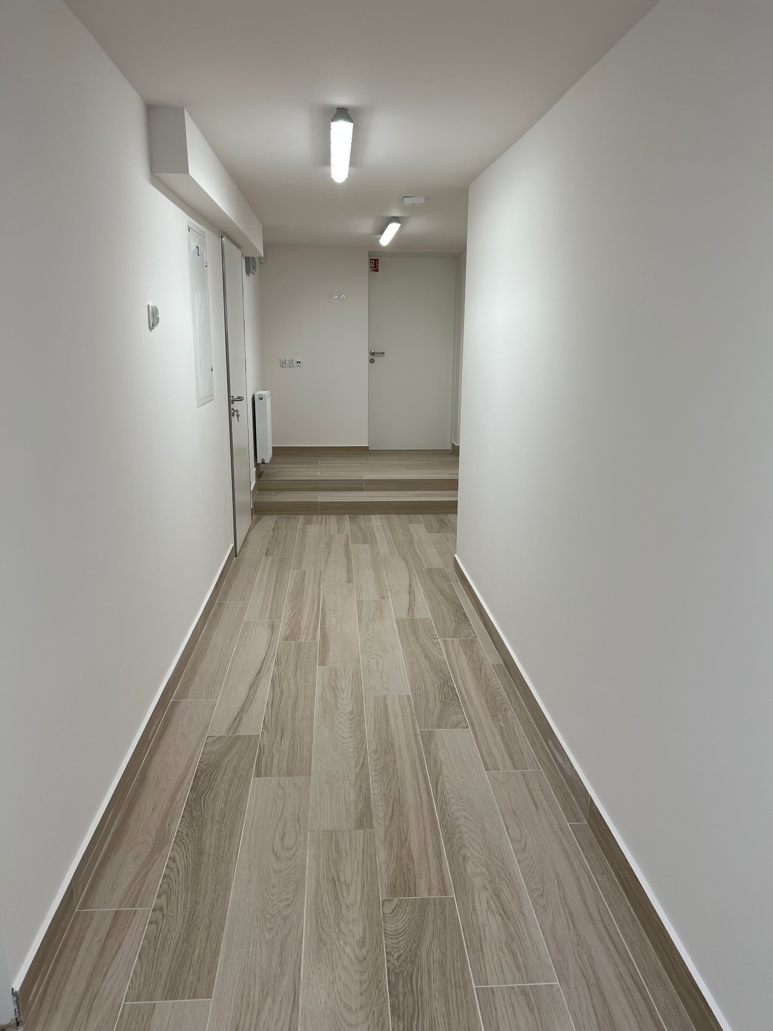Jasny korytarz z podłogą wyłożoną płytkami imitującymi drewno, świeżo pomalowane białe ściany i sufit z oświetleniem punktowym, widok na schody i drzwi w tle.