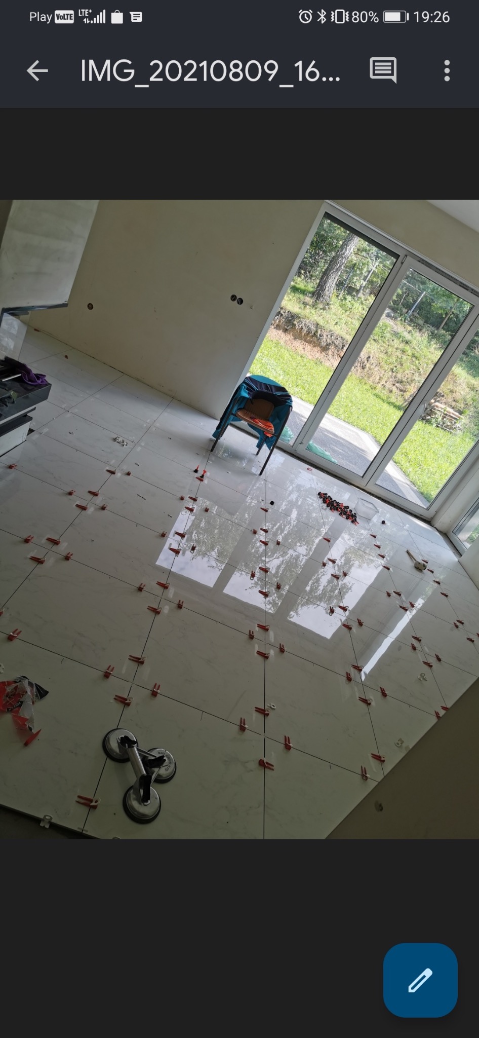 Błyszczące, białe płytki podłogowe układane z użyciem czerwonych krzyżyków dystansowych i przyssawki do przenoszenia płytek, widok na okno balkonowe i zielony ogród.