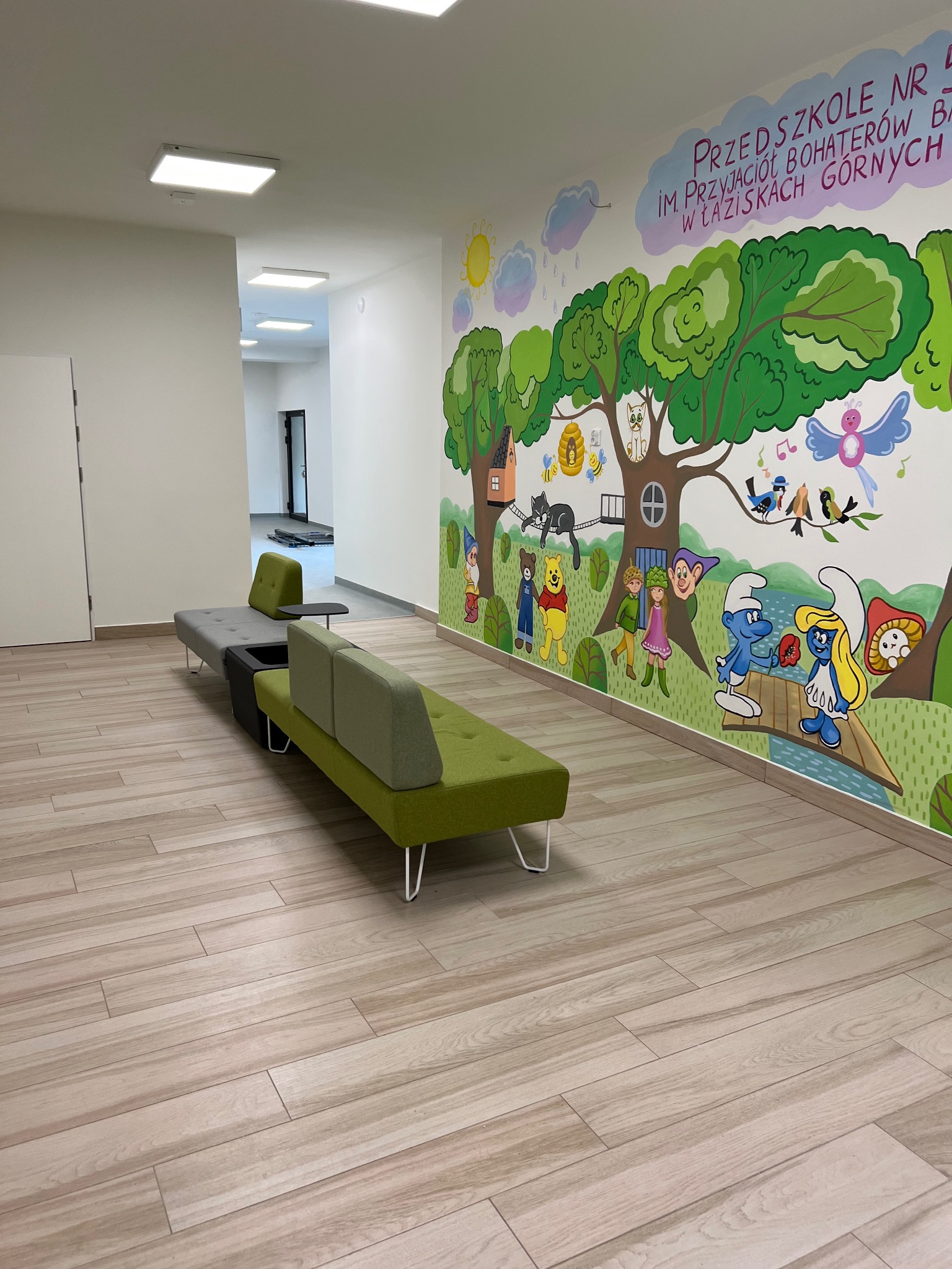 Korytarz przedszkola z drewnopodobną podłogą, nowoczesną sofą w odcieniach szarości i zieleni oraz muralem z postaciami z bajek na ścianie.