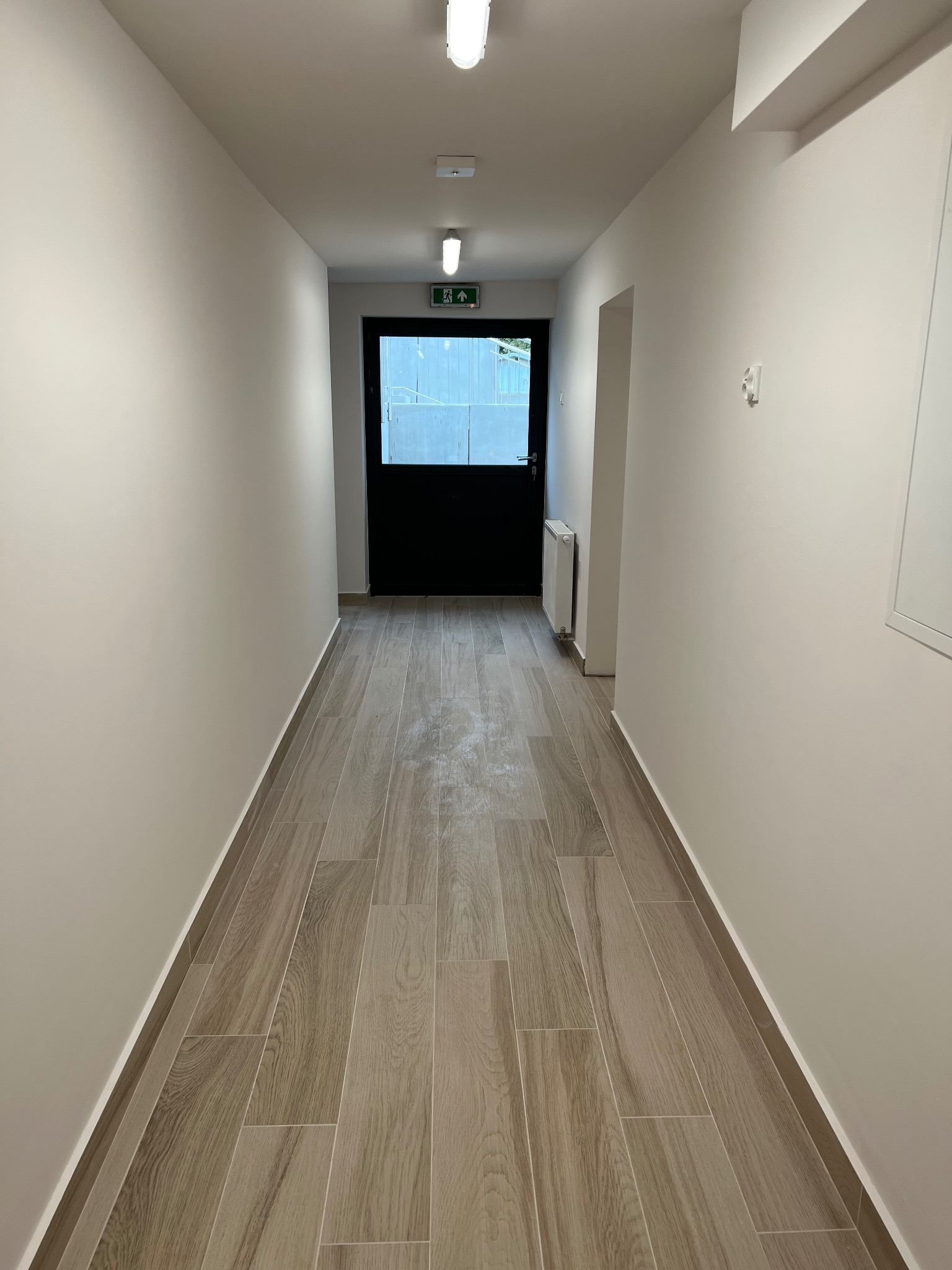 Korytarz z nowymi płytkami podłogowymi imitującymi drewno, świeżo pomalowane ściany w odcieniach bieli, czarne drzwi na końcu korytarza z widocznym znakiem ewakuacyjnym.