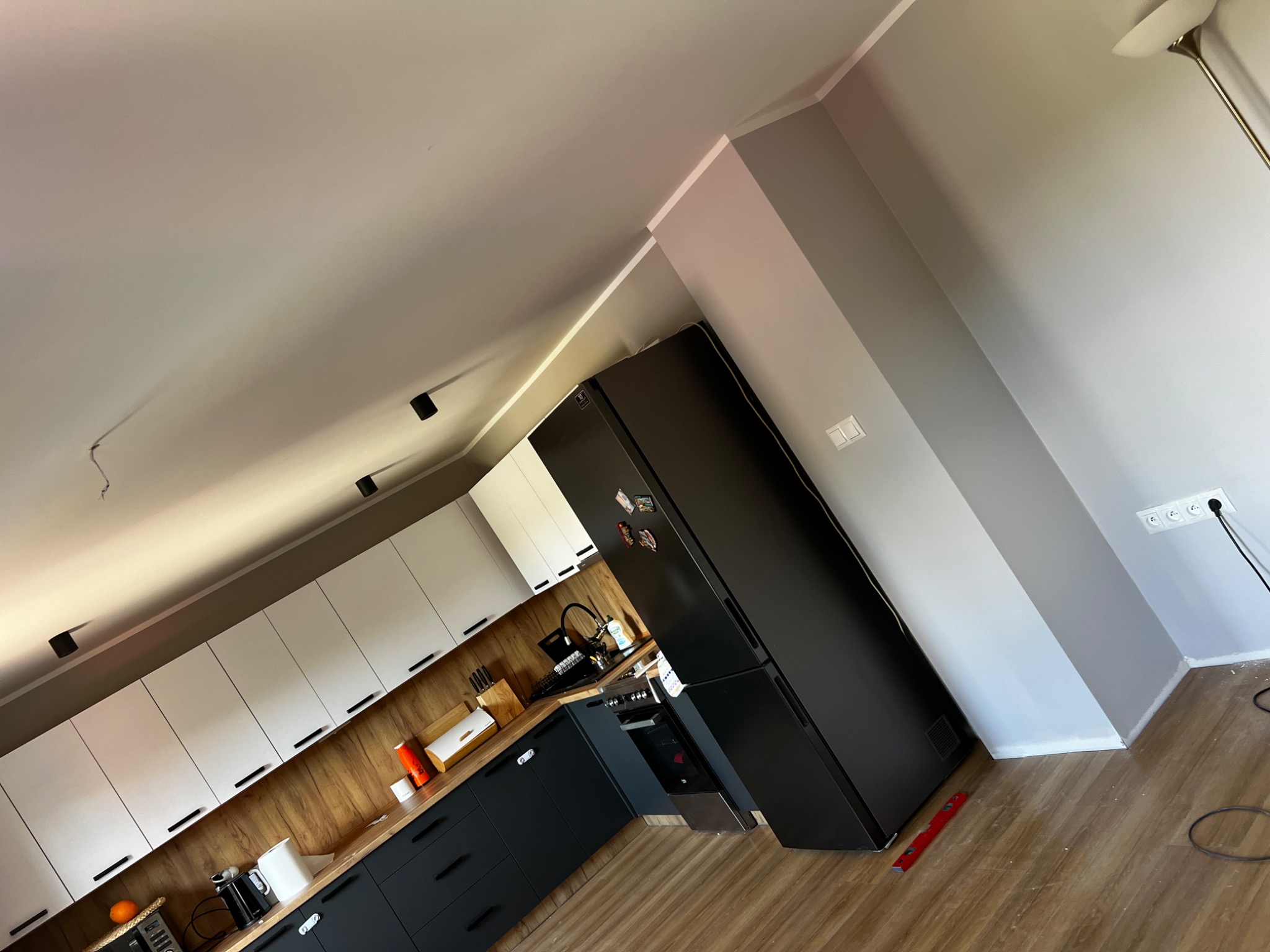 Nowoczesna kuchnia z białymi szafkami górnymi i grafitowymi dolnymi, drewnianym blatem i czarną lodówką; widoczny poziomica przy ścianie.