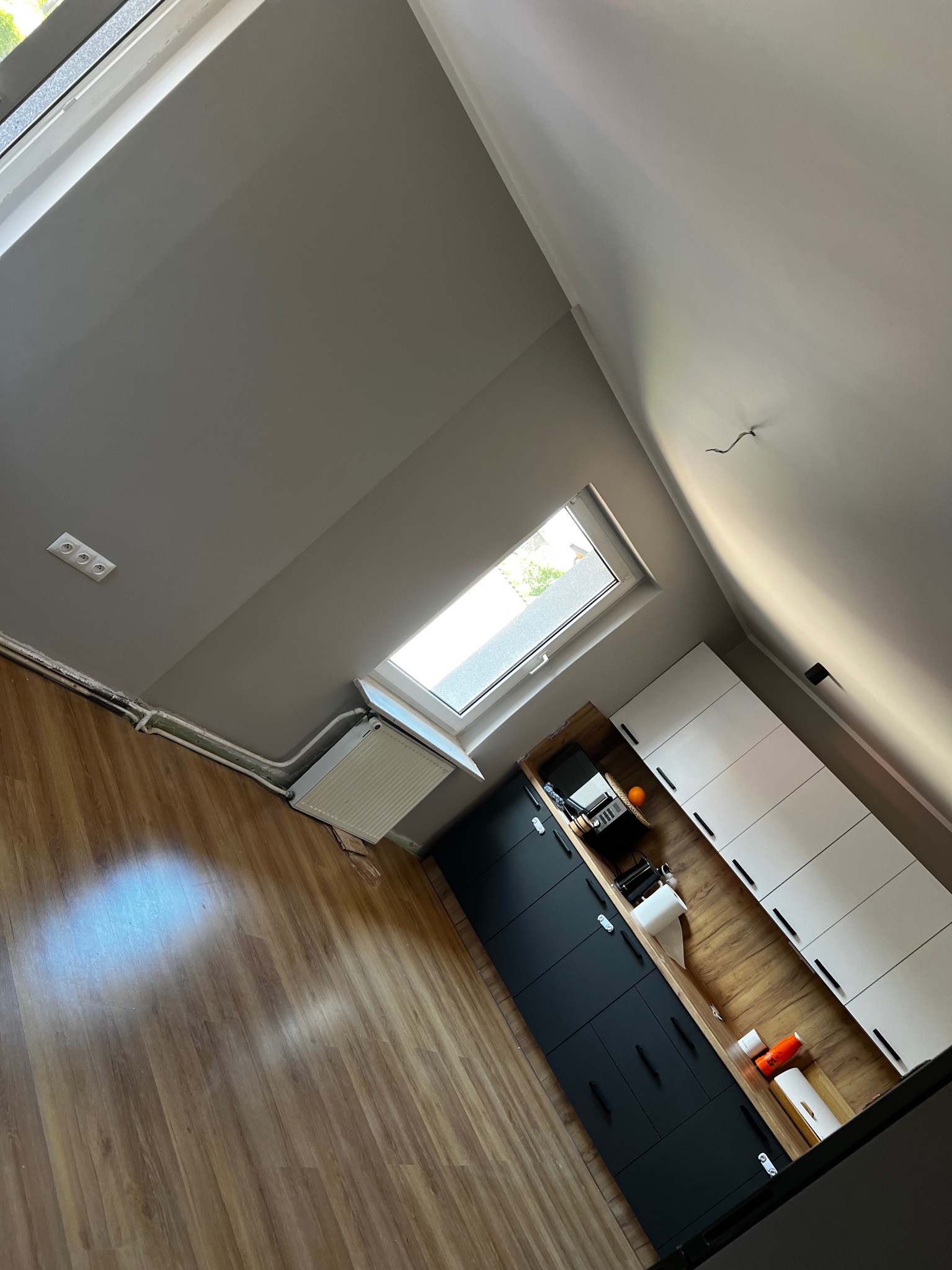 Szare, świeżo pomalowane poddasze z oknem dachowym, widoczne gładkie ściany, grzejnik, instalacja elektryczna, drewnopodobna podłoga oraz zabudowa kuchenna z czarnymi i białymi frontami.