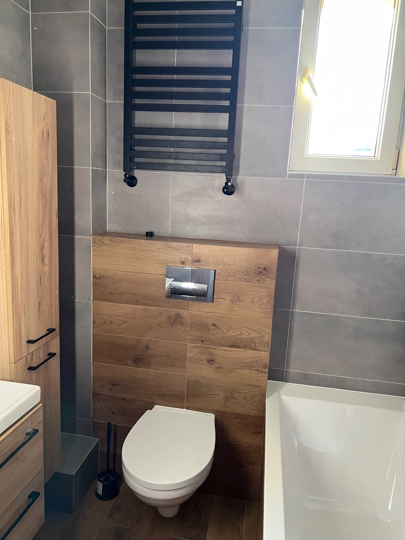 Nowoczesna łazienka z szarymi płytkami, drewnopodobną zabudową toalety, grzejnikiem drabinkowym i szafką z drewnianymi frontami.