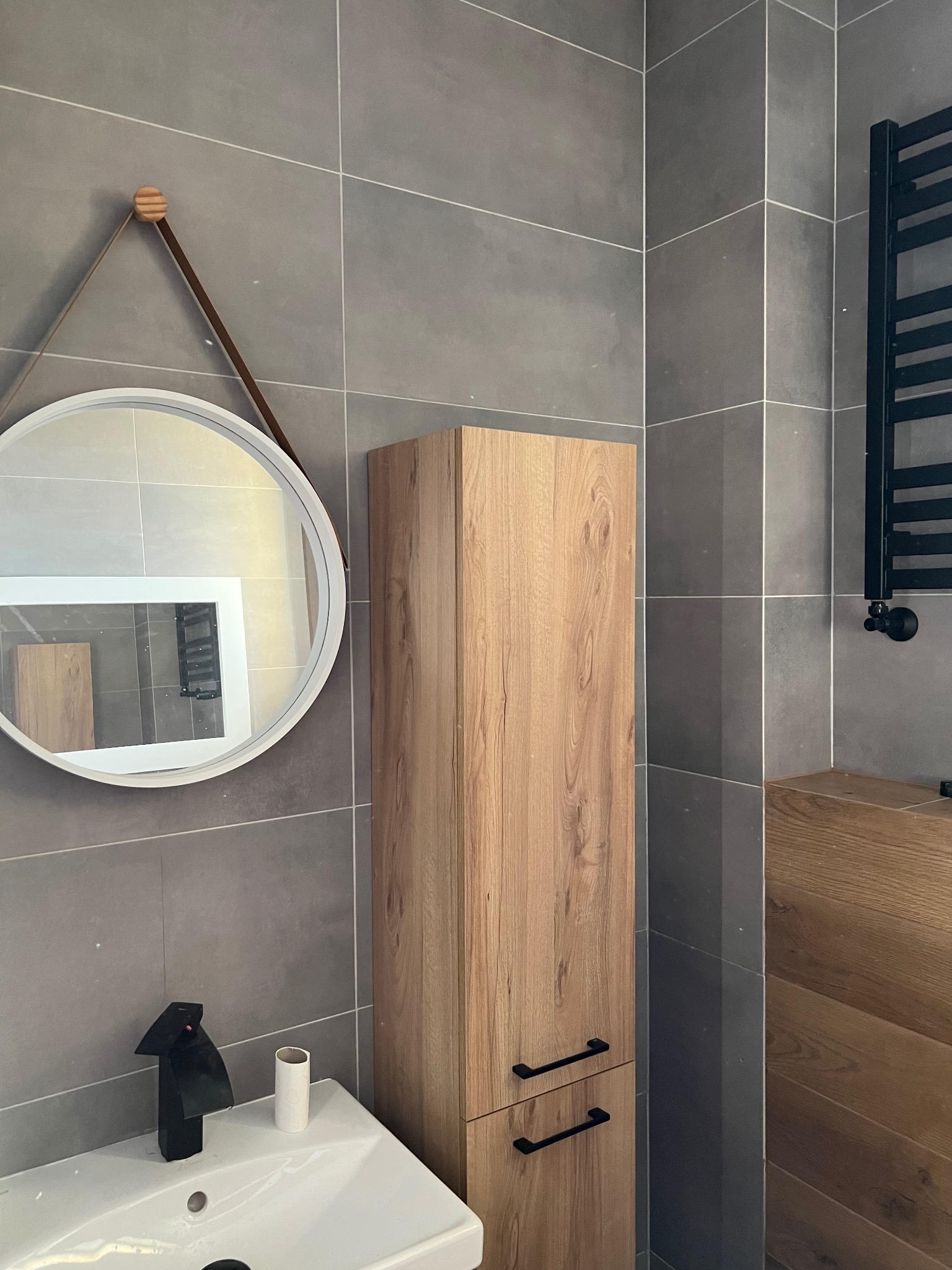 Nowoczesna łazienka z szarymi płytkami, okrągłym lustrem na skórzanym pasku, drewnianą szafką i czarną armaturą, widok na grzejnik drabinkowy.