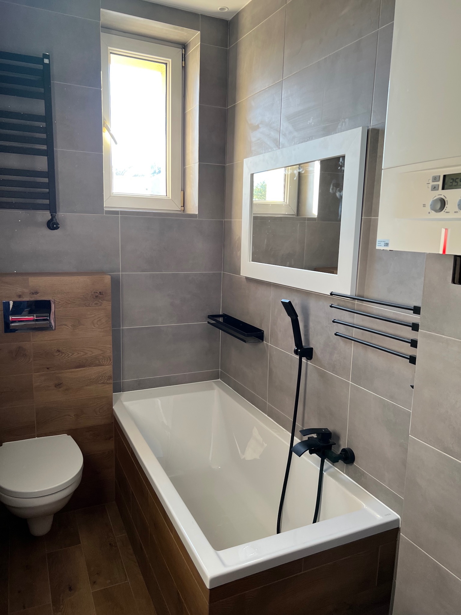 Nowoczesna łazienka w szaro-brązowych odcieniach z wanną obudowaną drewnopodobnymi panelami, wiszącą toaletą, grzejnikiem drabinkowym i czarną armaturą.