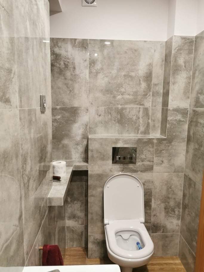 Wykończona łazienka z szarymi płytkami imitującymi kamień, wisząca toaleta z chromowanym przyciskiem spłukiwania, półka z rolką papieru toaletowego i ręcznikiem.