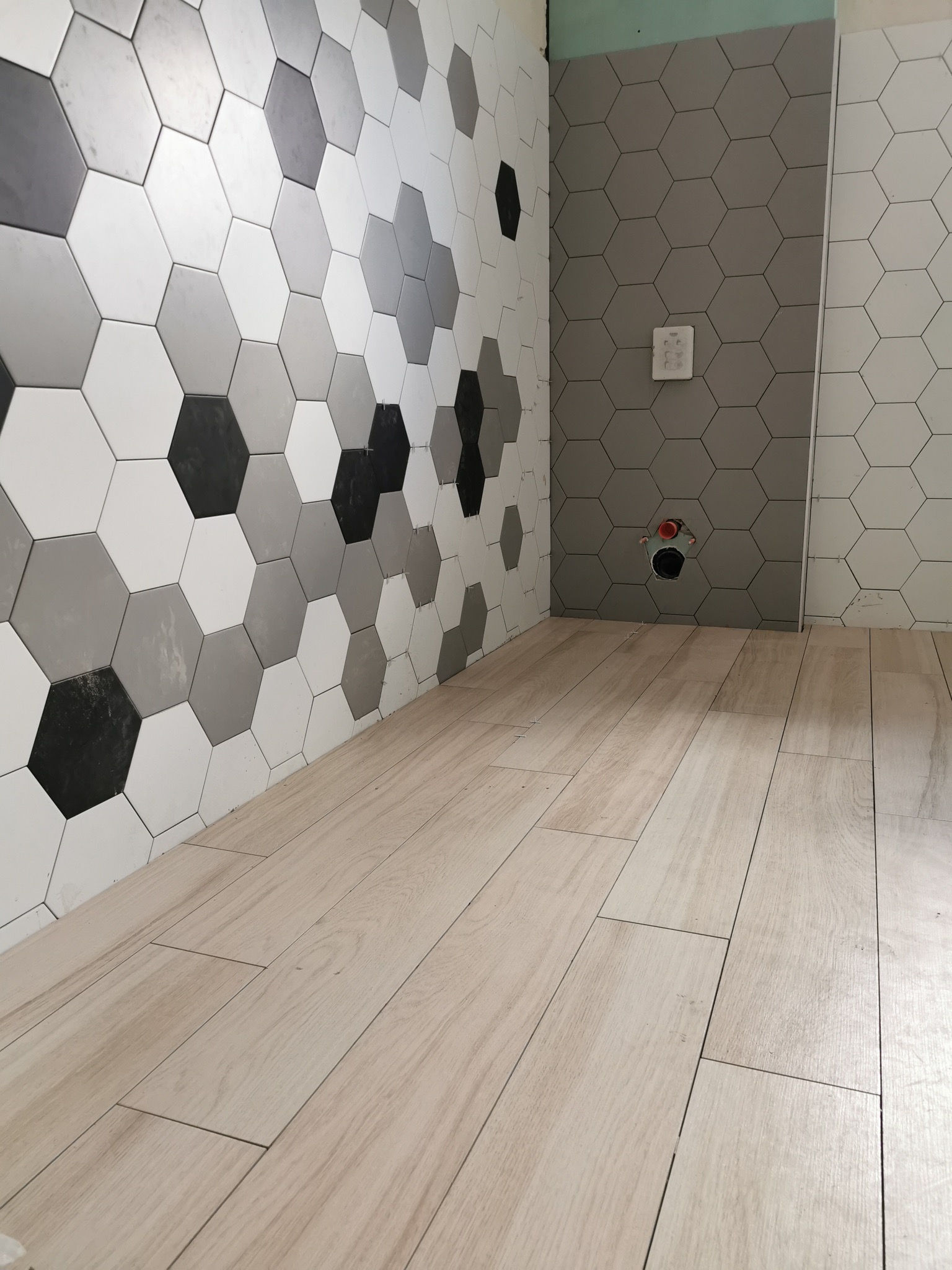 Ściana wyłożona heksagonalnymi płytkami w odcieniach szarości, bieli i czerni, zestawiona z podłogą z płytek imitujących jasne drewno w trakcie montażu w łazience.
