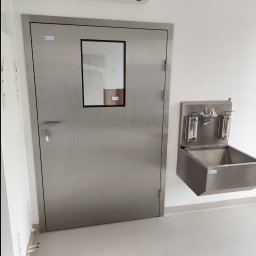 Piotr Żwirblis SERVIS - Stalowe drzwi z okienkiem i umywalka ze stali nierdzewnej w pomieszczeniu o sterylnych warunkach, prawdopodobnie w placówce medycznej w Płośnicy.