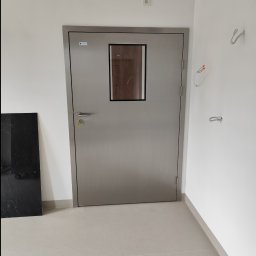 Piotr Żwirblis SERVIS - Stalowe drzwi wewnętrzne z małym okienkiem, widoczne przewody elektryczne na ścianie, obok czarna płyta marmurowa.