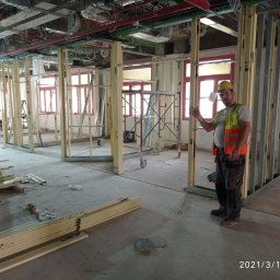 Piotr Żwirblis SERVIS - Pracownik w żółtym kasku i pomarańczowej kamizelce prezentuje szkielet ścian działowych z profili metalowych w trakcie remontu mieszkania, widoczne instalacje pod sufitem i rusztowanie.