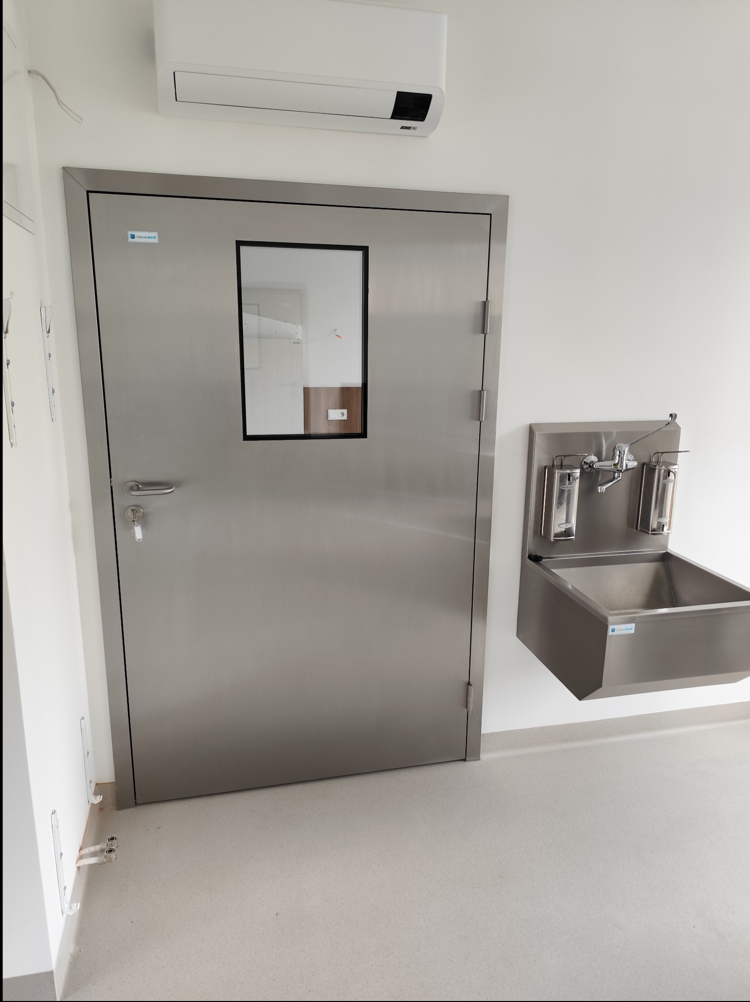 Stalowe drzwi z okienkiem i umywalka ze stali nierdzewnej w pomieszczeniu o sterylnych warunkach, prawdopodobnie w placówce medycznej w Płośnicy.