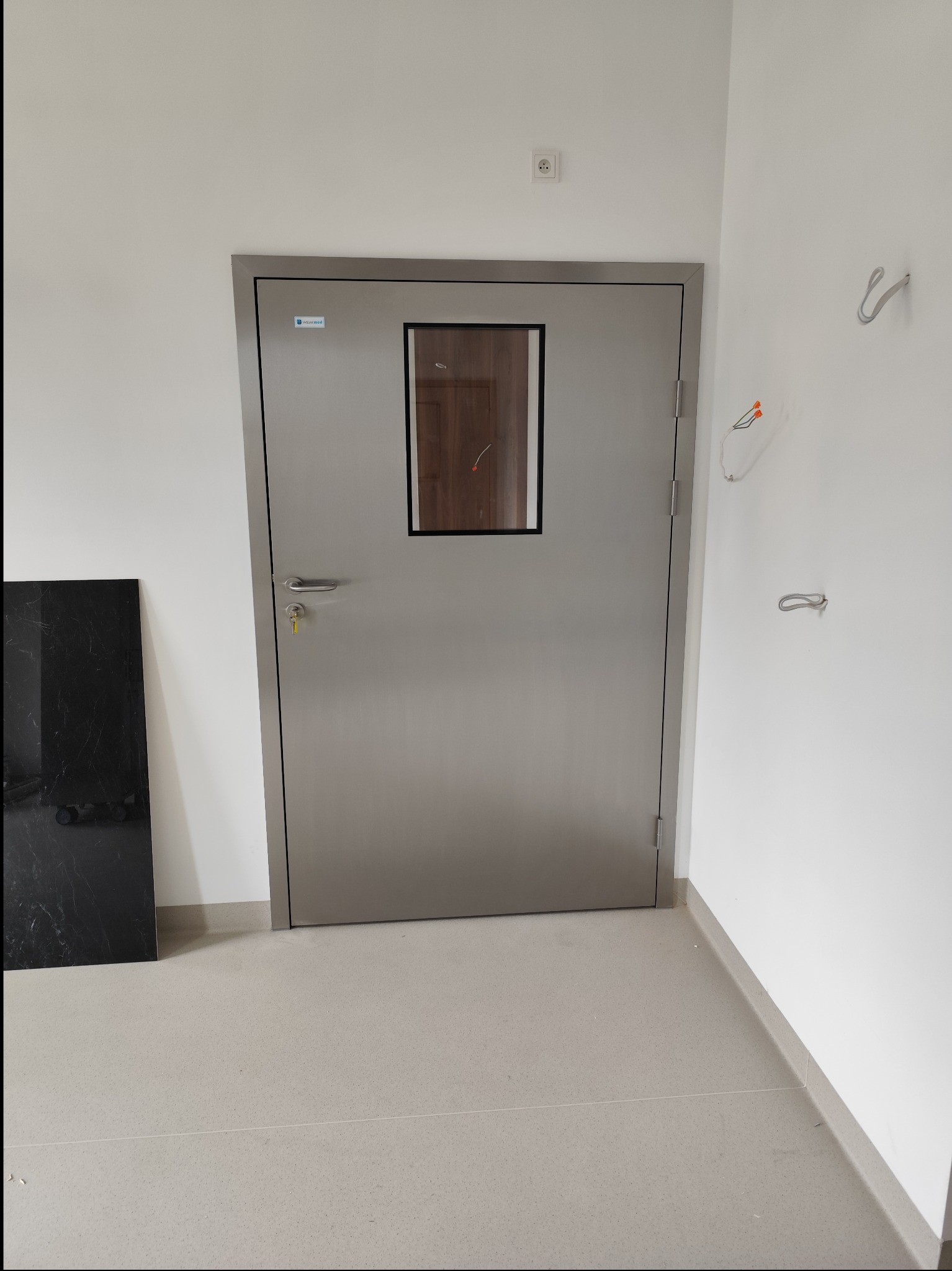 Stalowe drzwi wewnętrzne z małym okienkiem, widoczne przewody elektryczne na ścianie, obok czarna płyta marmurowa.