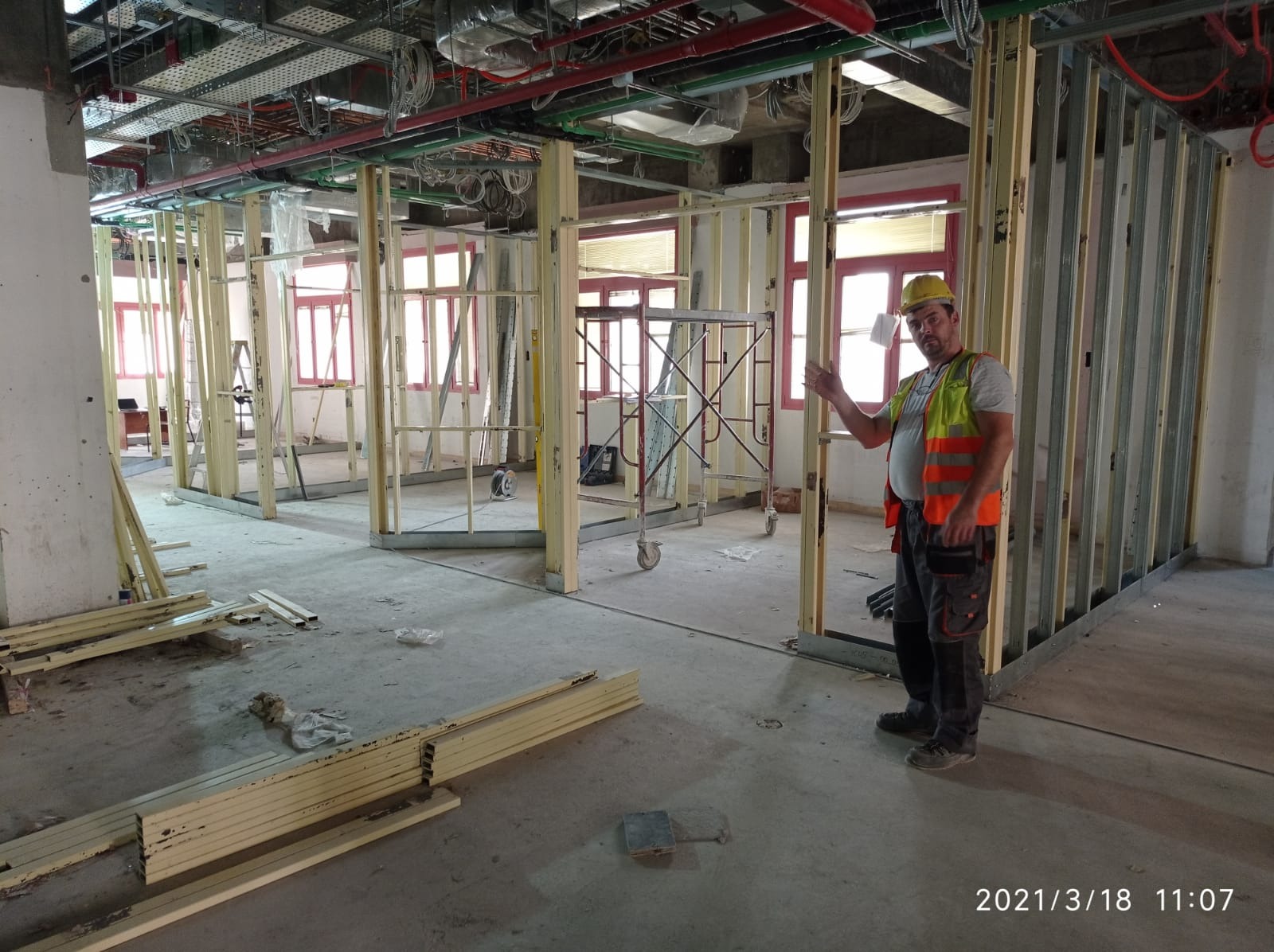 Pracownik w żółtym kasku i pomarańczowej kamizelce prezentuje szkielet ścian działowych z profili metalowych w trakcie remontu mieszkania, widoczne instalacje pod sufitem i rusztowanie.